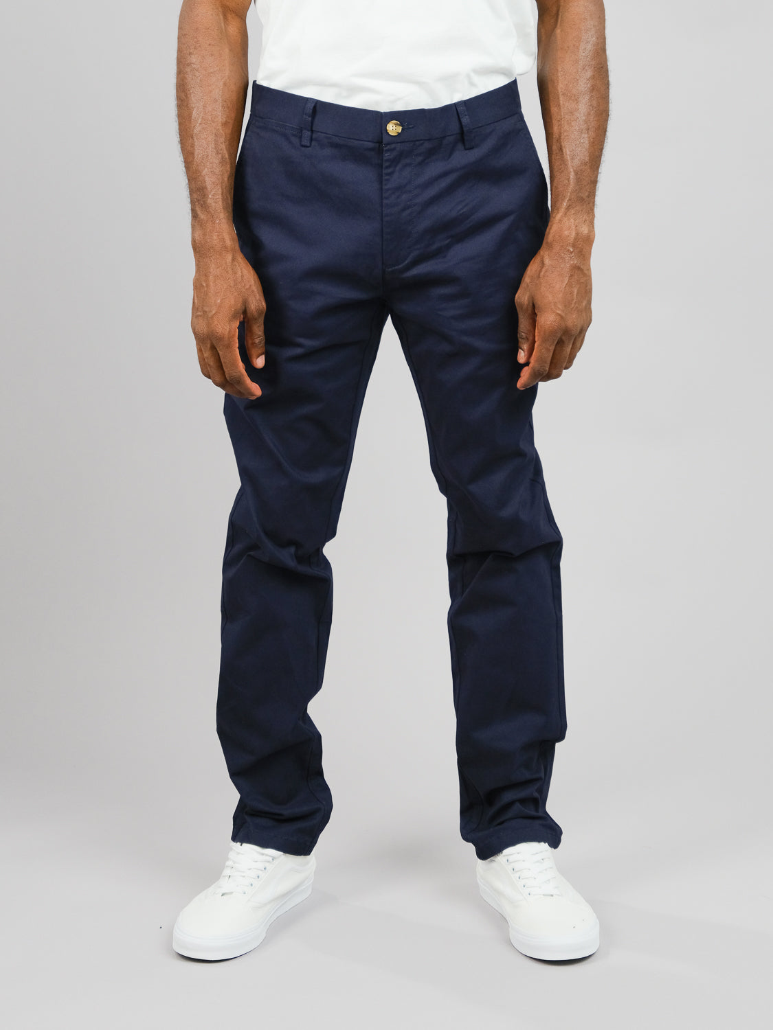 A person wearing a white shirt and ID Menswears ID Brooklyn Chino trousers in a regular slim fit, stands against a plain gray background. Their hands rest by their sides, complemented by white sneakers. The image is cropped at the shoulders.