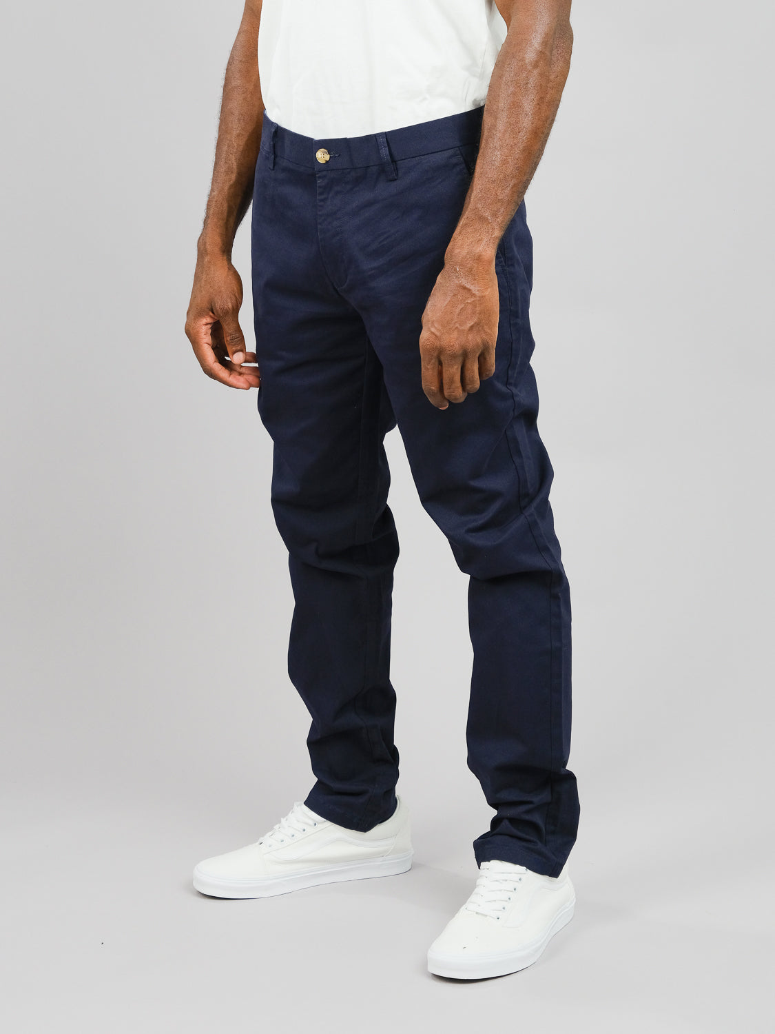 A person models the white ID Brooklyn Chino from ID Menswear and navy blue pants with white sneakers, standing against a plain gray background. Their hands rest by their sides, emphasizing the regular slim fit of this cotton twill outfit.
