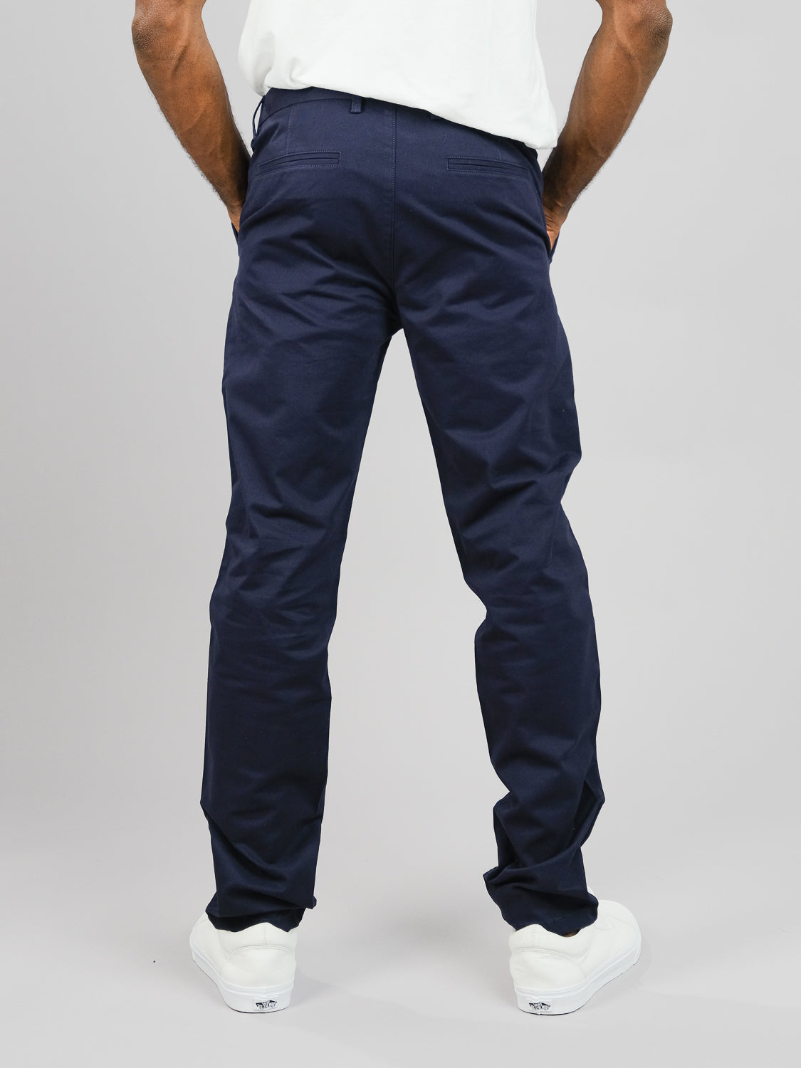 Someone stands against a plain background, wearing ID Menswears navy blue ID Brooklyn Chino in a regular slim fit crafted from cotton twill, paired with white sneakers. The image highlights the lower body showcasing the back view of the pants.