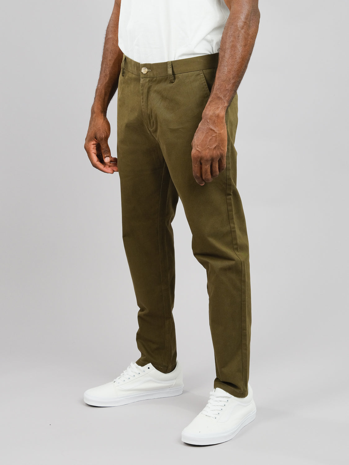 Against a plain background, a person showcases the lower half of their body in olive green ID Brooklyn Chinos from ID Menswear, made of cotton twill. Theyre paired with a white T-shirt and sneakers, subtly elevating this casual look.