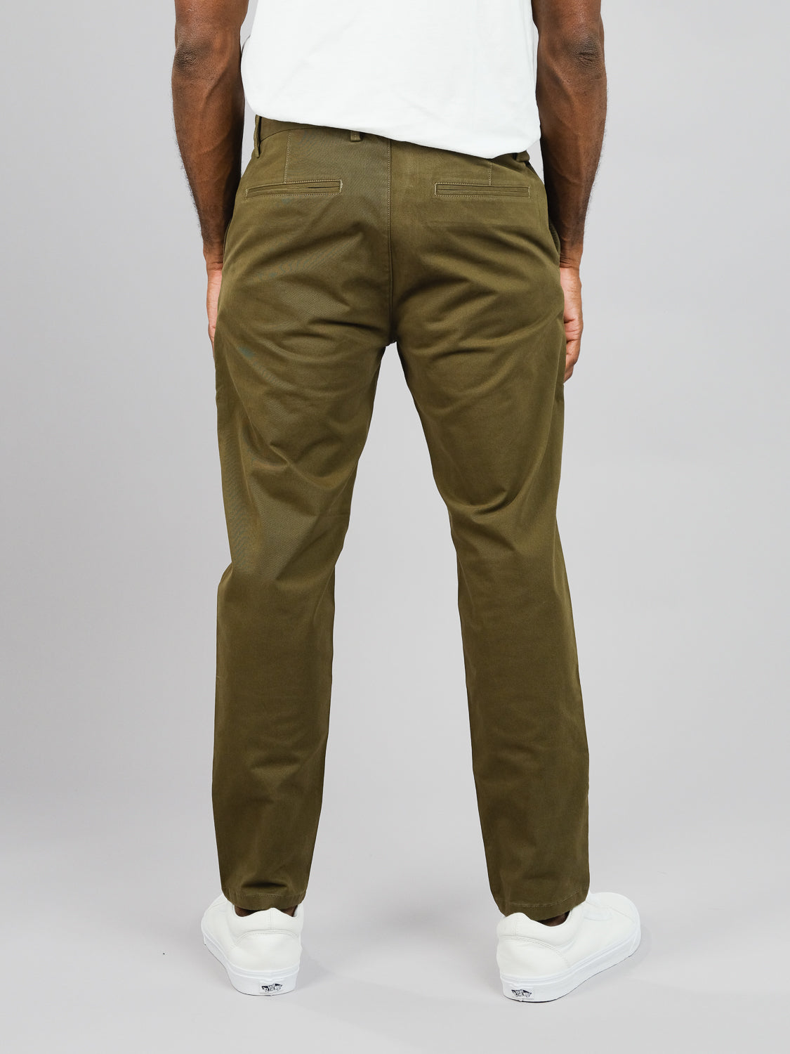 A person wearing olive green ID Brooklyn Chino pants by ID Menswear and white sneakers stands against a plain gray background. The shot highlights the regular slim fit and cotton twill fabric from behind, with hands at their sides.