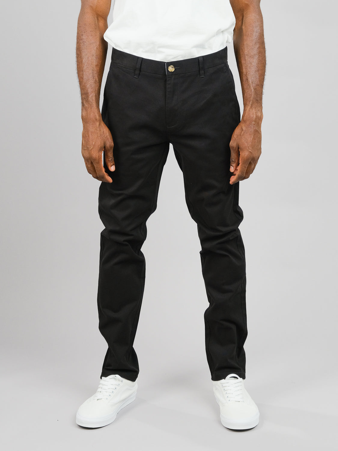 A person in black ID Menswear ID Brooklyn Chino pants, a white shirt, and white sneakers poses against a gray background, highlighting the regular slim fit of the cotton twill chinos along with the matching shoes.