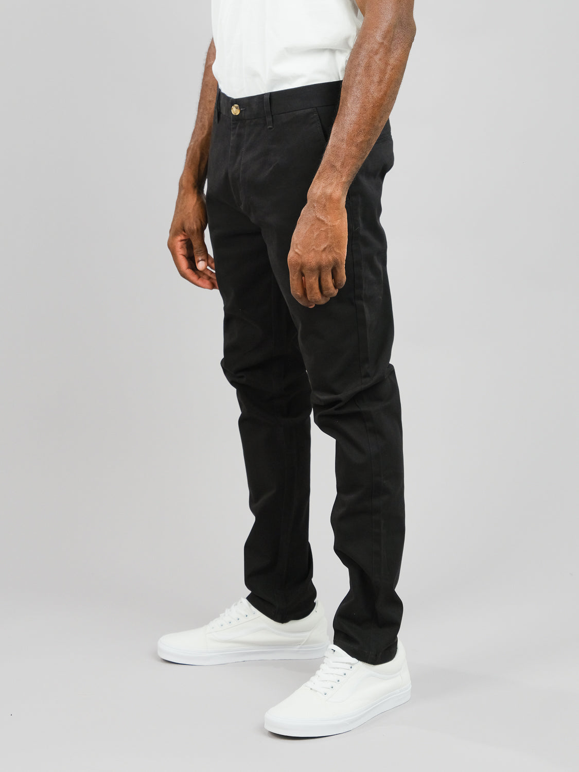 Against a plain gray backdrop, a person in a white shirt and sneakers with black pants embodies the relaxed elegance of ID Brooklyn Chino by ID Menswear, crafted from soft cotton twill with hands resting loosely by their sides.