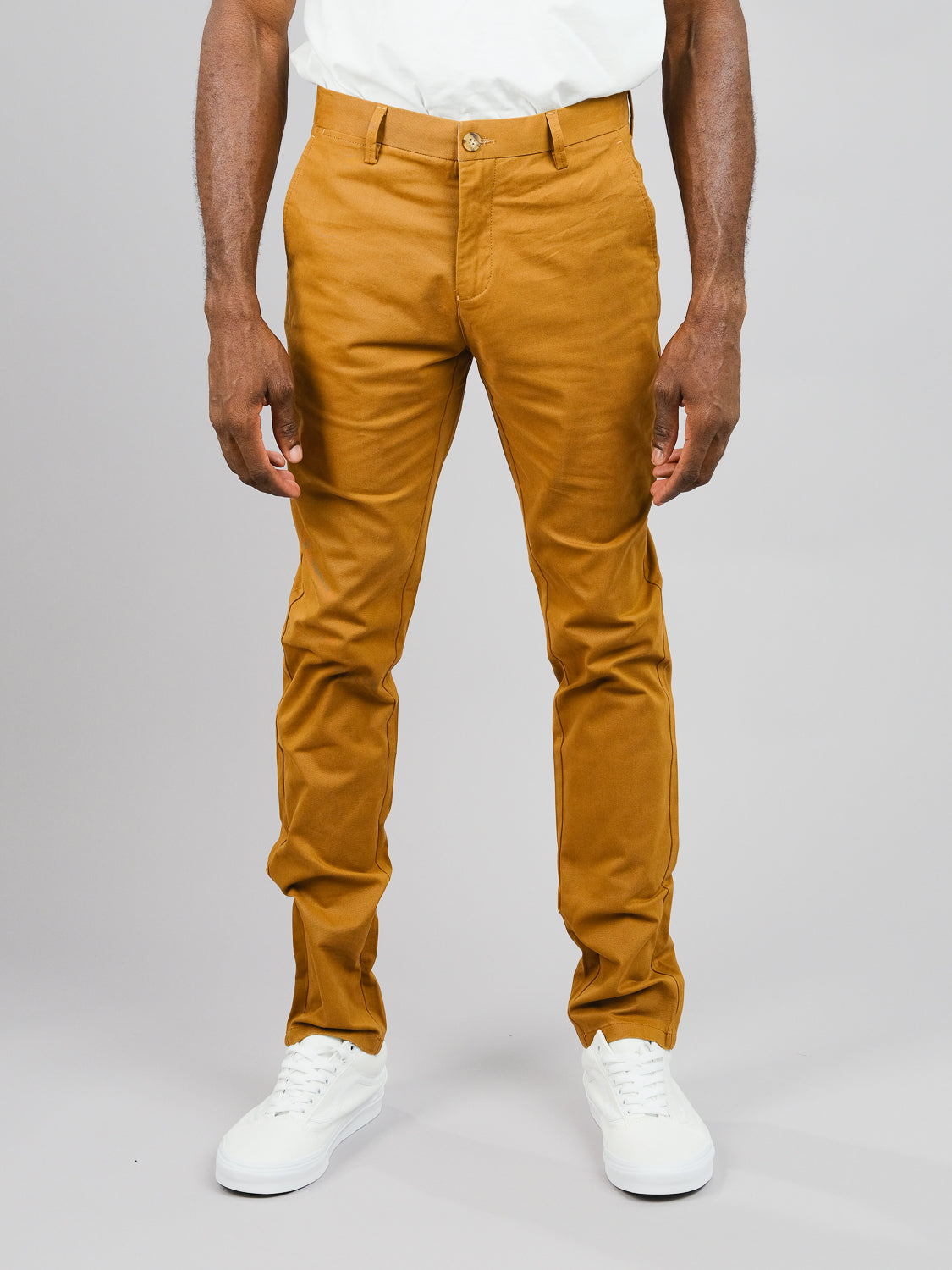 A man stands against a plain background, showcasing the ID Menswear ID Brooklyn Chino in mustard cotton twill. The regular slim fit chinos pair well with his white sneakers and shirt, with hands at his sides. The image highlights the lower half of his body.