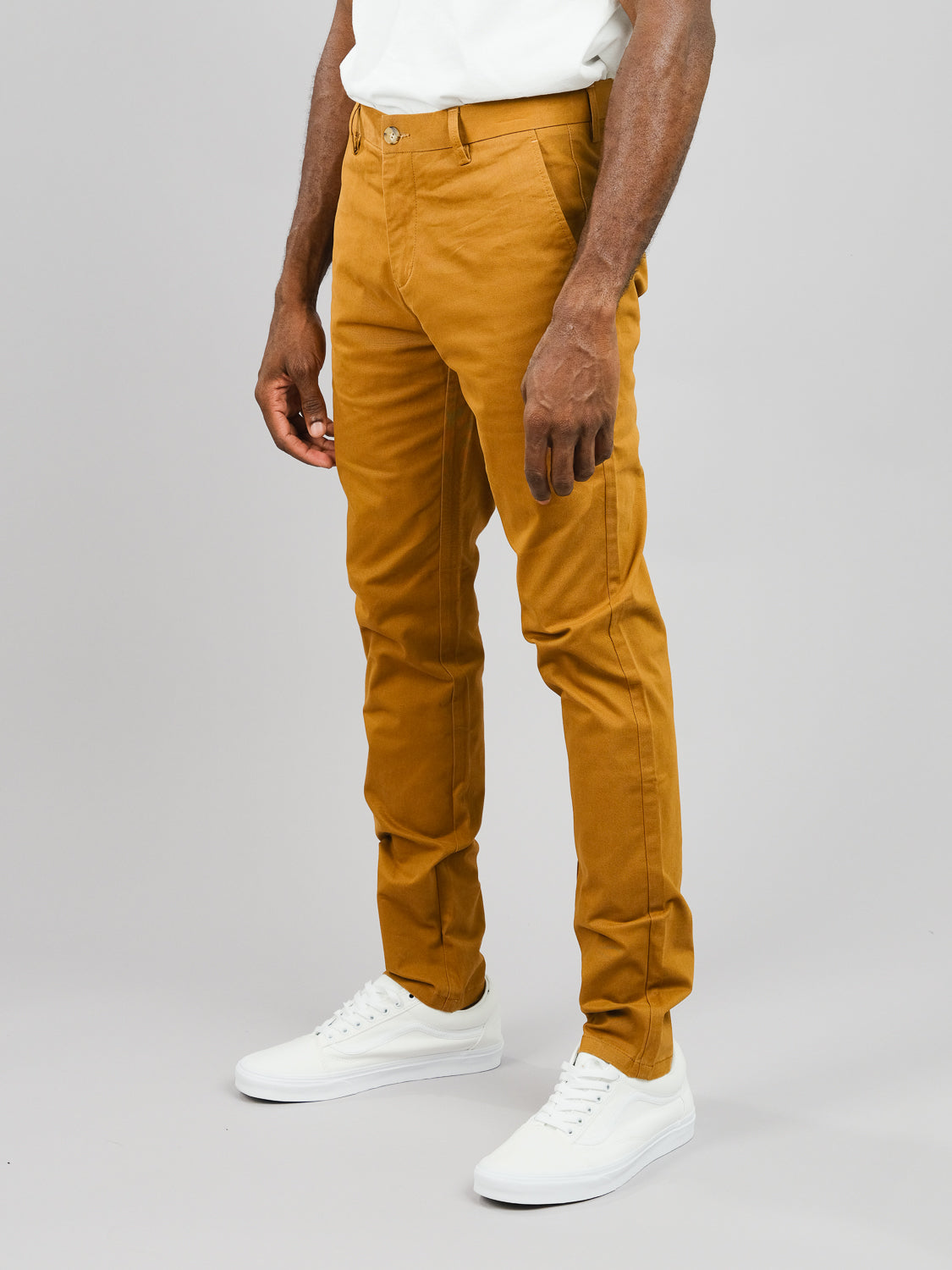 A person in mustard ID Menswears Brooklyn Chino, regular slim fit, with white sneakers. Their upper body is partially visible, arms relaxed by their sides against a plain light gray background.