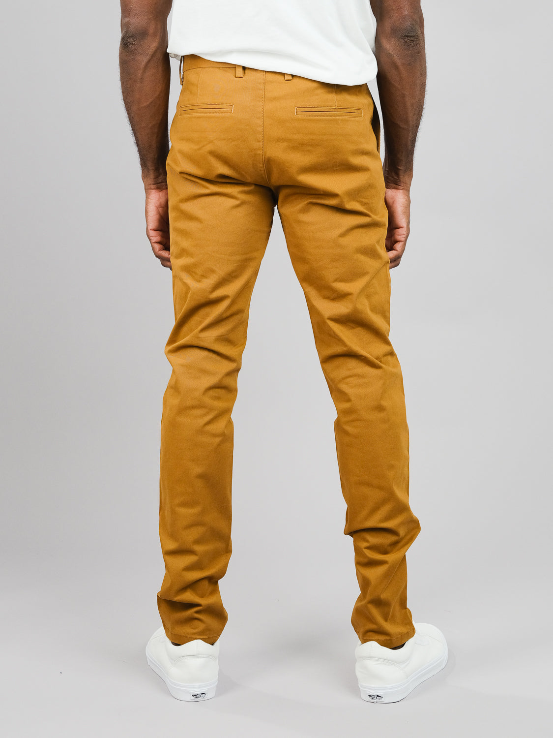 A person is seen from behind wearing a mustard ID Brooklyn Chino by ID Menswear in a regular slim fit paired with white sneakers, set against a plain gray backdrop.