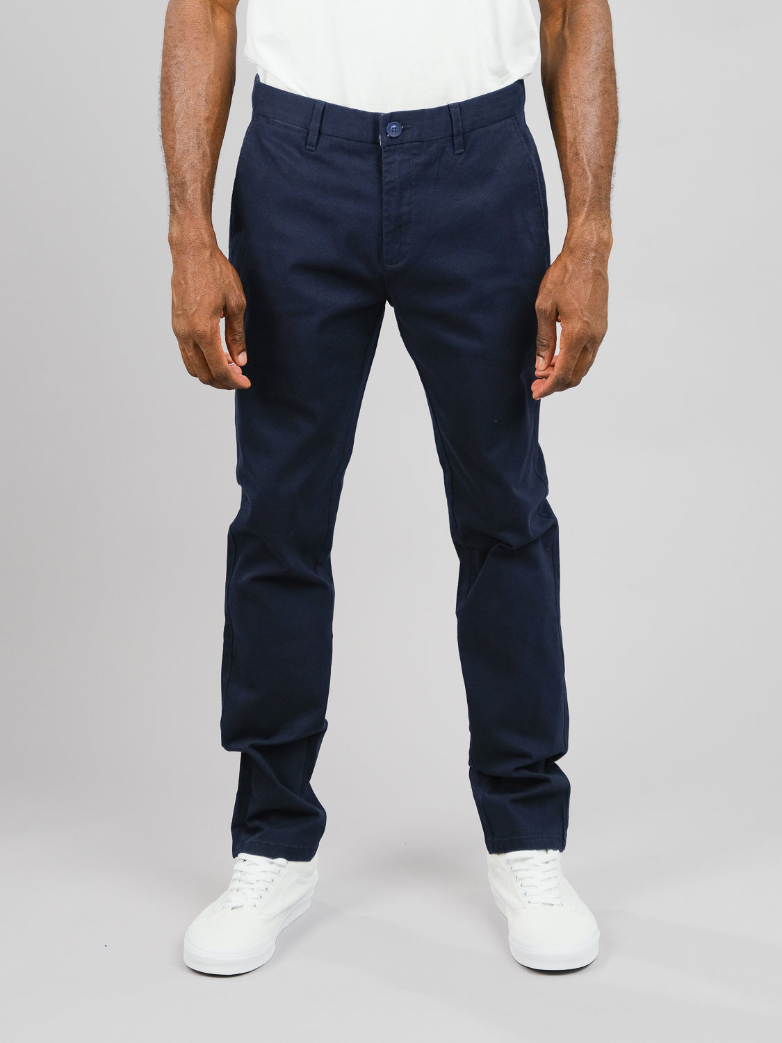 Against a light gray background, a person showcases versatile style in ID Menswears Wythe Twill Stretch Chino, slim-fit navy blue cotton twill chinos, paired with white sneakers. The upper body is cropped above the mid-chest, emphasizing casual elegance.