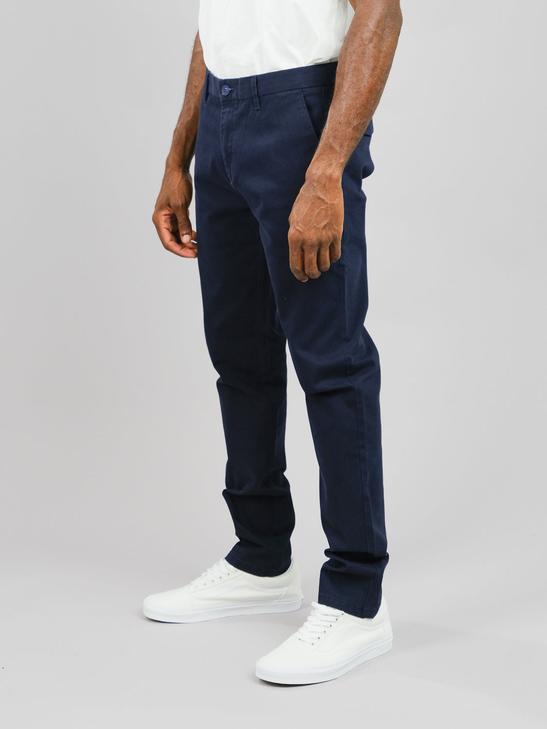 A person wears ID Menswears Wythe Twill Stretch Chino, a pair of slim-fit dark blue pants, paired with a white shirt and sneakers, standing against a light gray background. The focus is on their lower half, highlighting the neat style perfect for casual occasions.