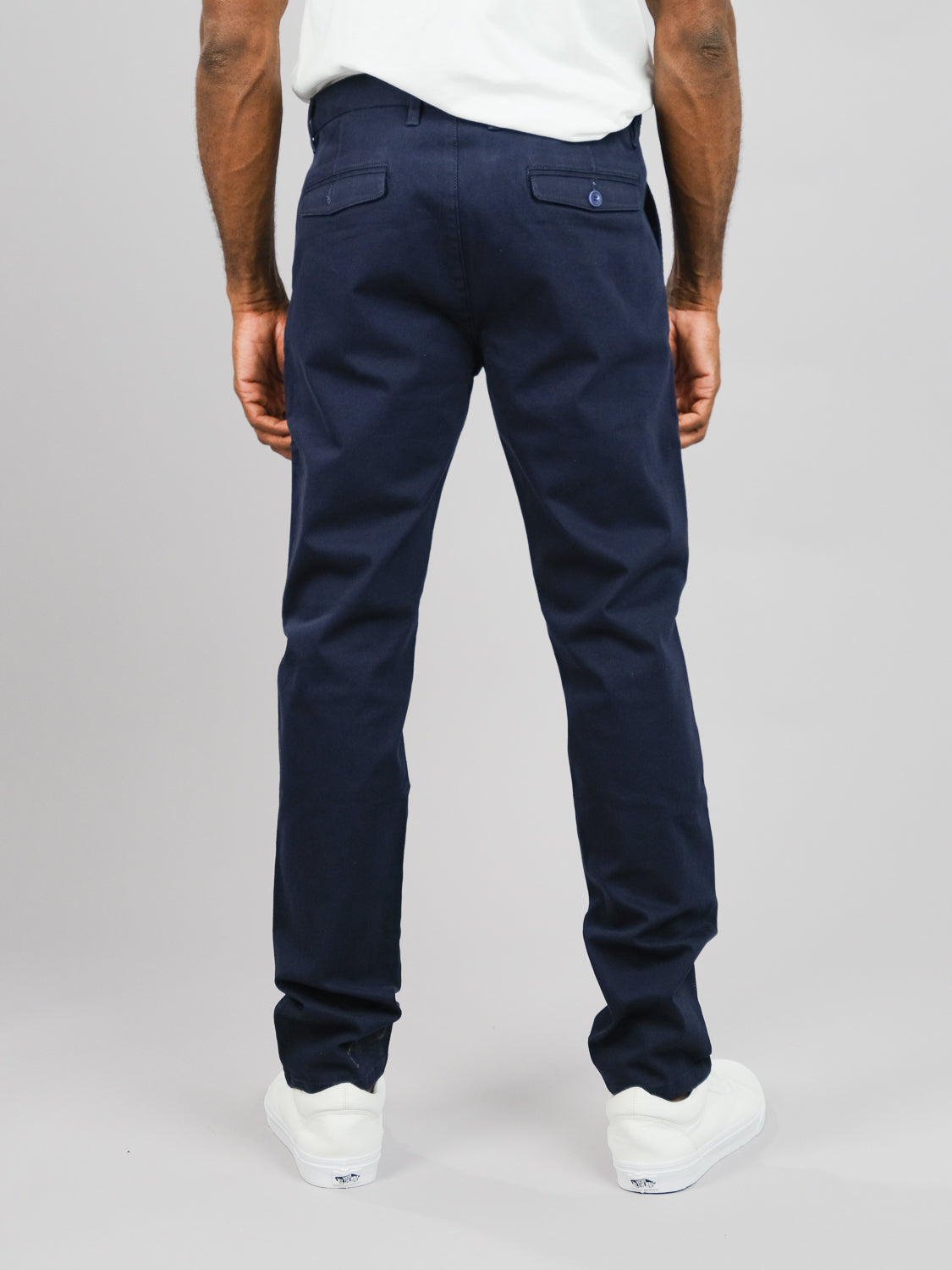 Wearing ID Menswears Wythe Twill Stretch Chino, a navy blue slim-fit pant with back pockets, the person pairs it with a white shirt and sneakers, perfect for casual settings. They stand before a plain gray background.