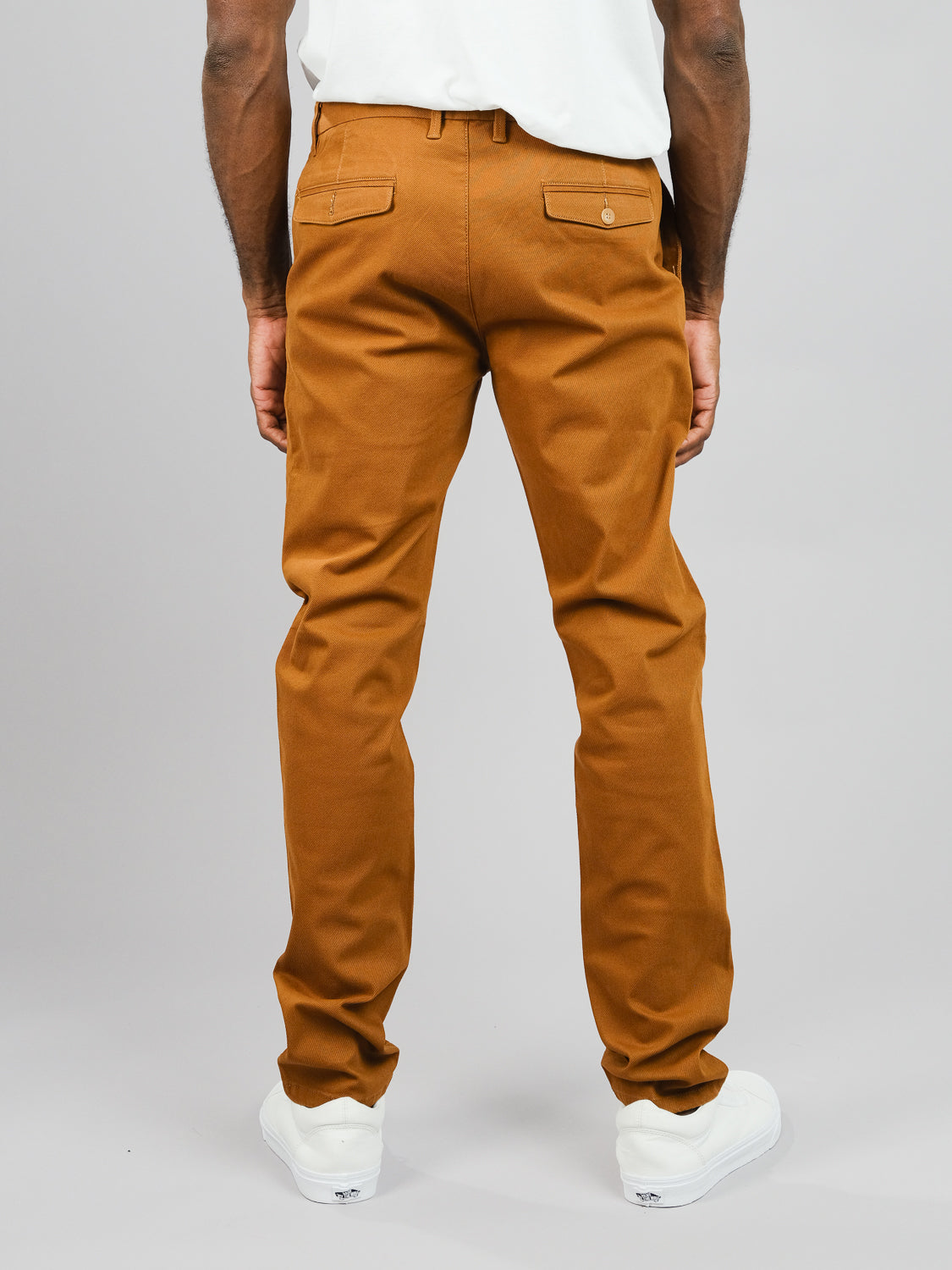A person wearing ID Menswears Wythe Twill Stretch Chino pants in brown and a white shirt is seen from behind. The chinos, with buttoned back pockets, suit casual outings, paired seamlessly with white sneakers against a gray backdrop.