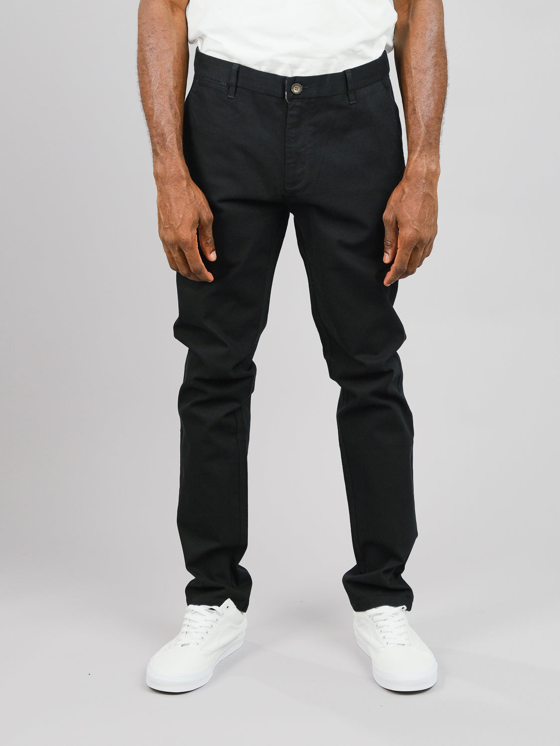 Against a gray background, a person models slim-fit Wythe Twill Stretch Chino by ID Menswear. The black cotton twill pants, perfect for casual occasions, feature a button closure and visible seams. White sneakers complete the look as their hands rest by their sides with legs and lower torso in view.