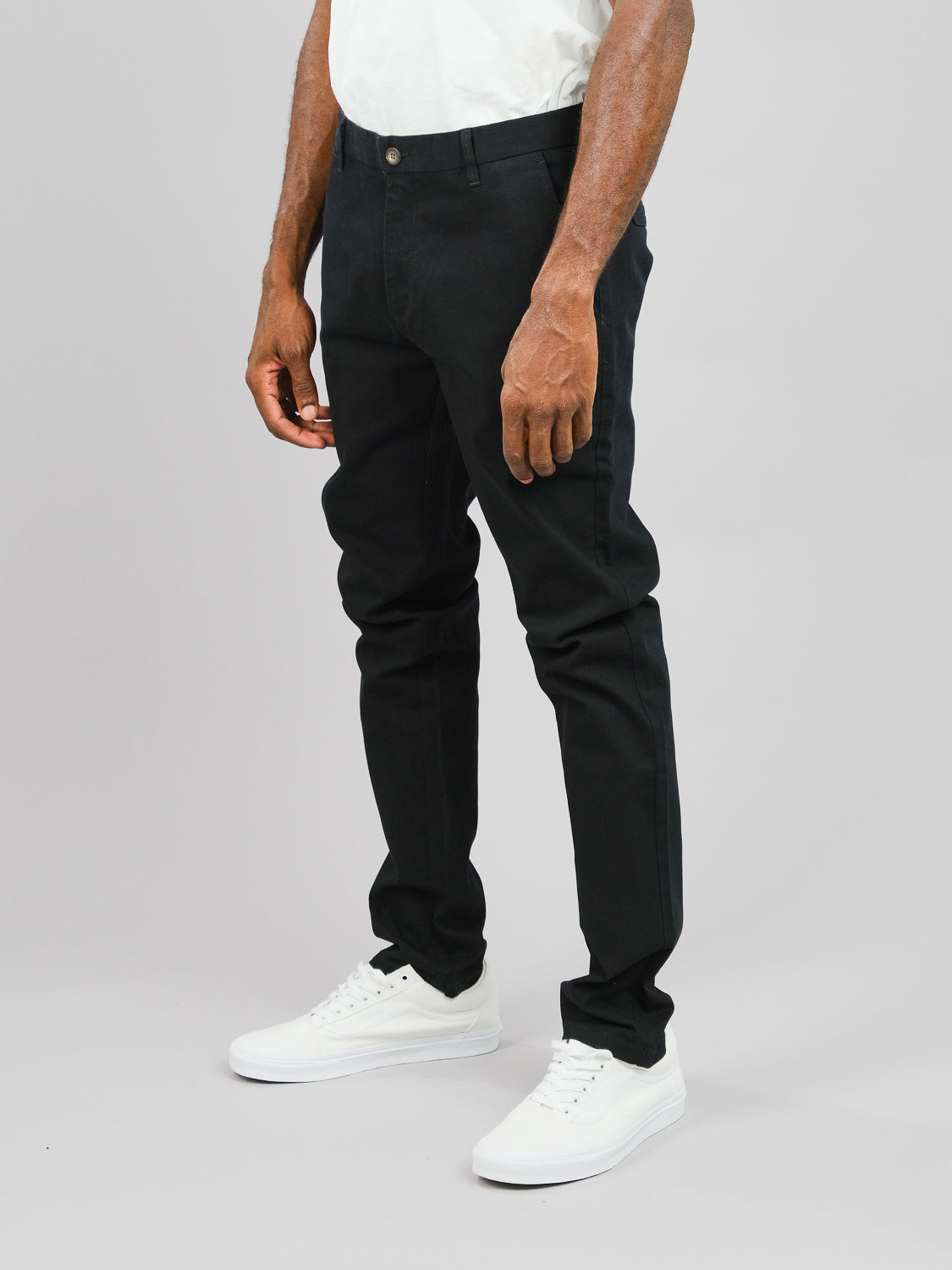 Clad in ID Menswears Wythe Twill Stretch Chino, featuring slim-fit black cotton twill fabric, a person stands against a plain gray backdrop. With white sneakers and hands resting by their side, this look is ideal for casual occasions, focusing on the sleek design of the chinos torso and leg-fitting style.