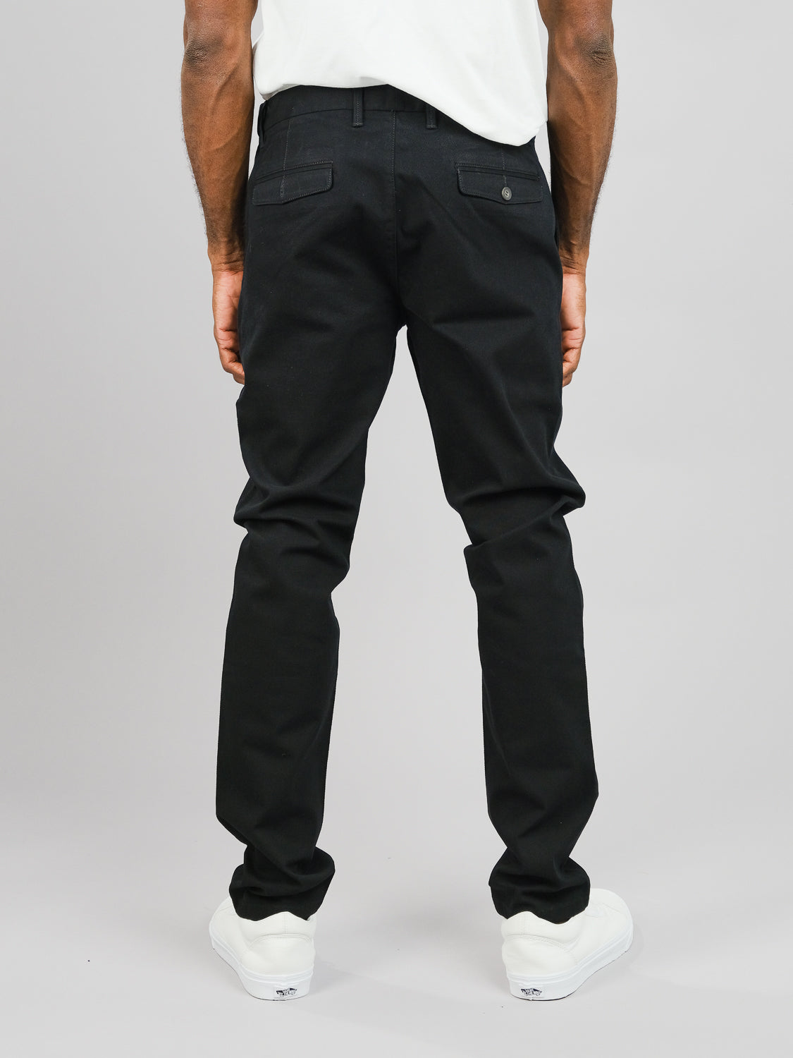 A person is shown from behind, wearing ID Menswears Wythe Twill Stretch Chino and white sneakers against a gray backdrop. The photo highlights the pants slim-fit style, perfect for casual events.