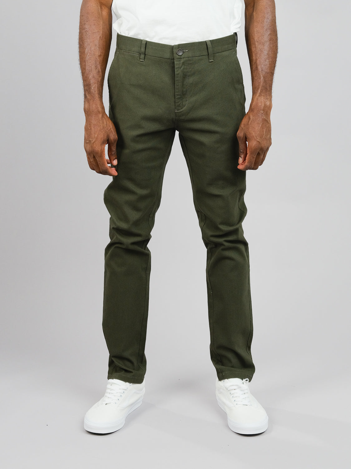 Sporting the Wythe Twill Stretch Chino in olive green from ID Menswear, a figure stands against a gray backdrop wearing cotton twill slim-fit pants with button and zip closure, complemented by white sneakers and a white t-shirt. Only the lower half is visible, ideal for casual outings.