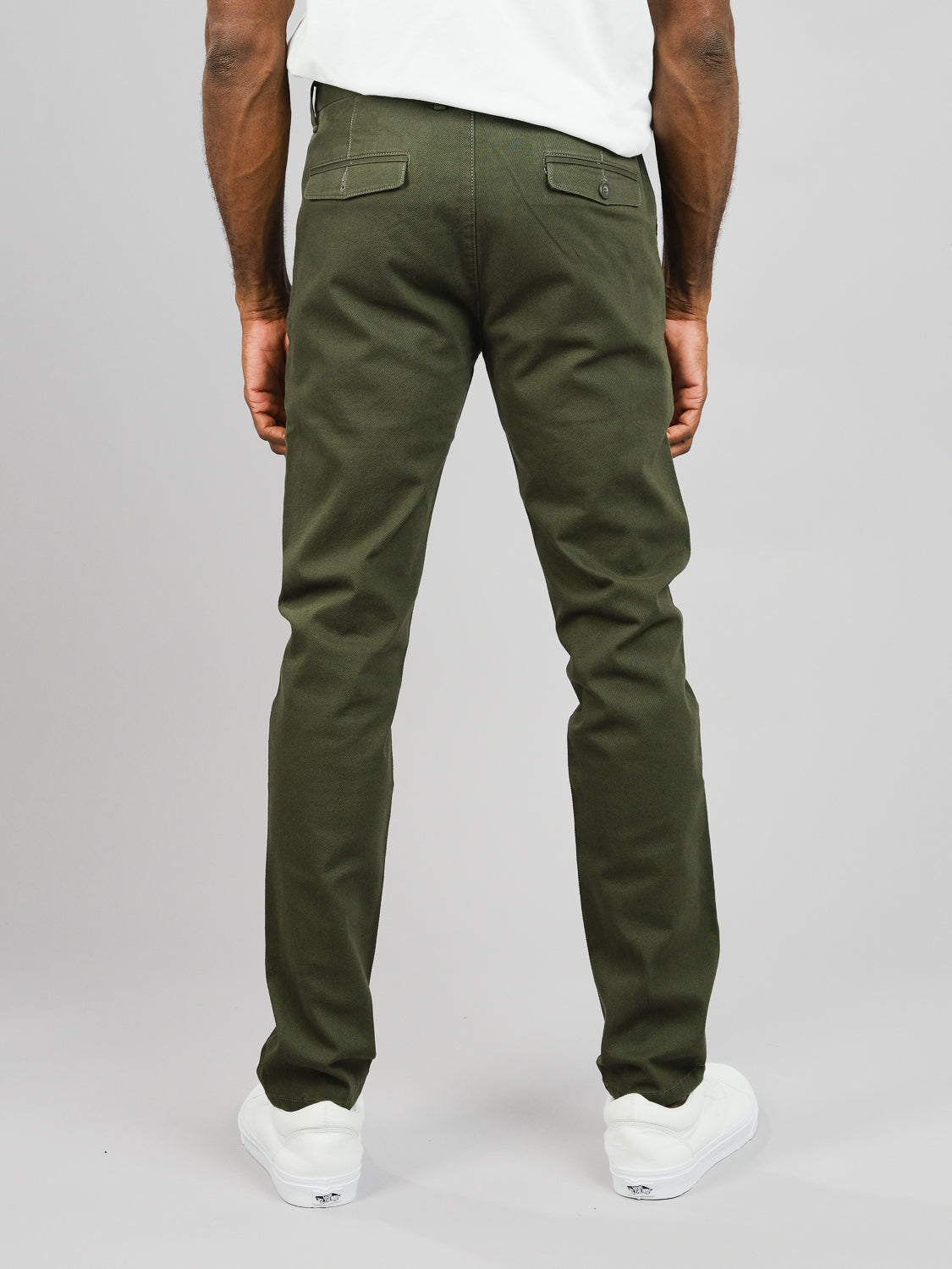 A person wears ID Menswears Wythe Twill Stretch Chino in olive green, featuring slim-fit cotton twill and buttoned back pockets, paired with a white shirt and sneakers. The outfit suits casual occasions against a plain, light gray background.