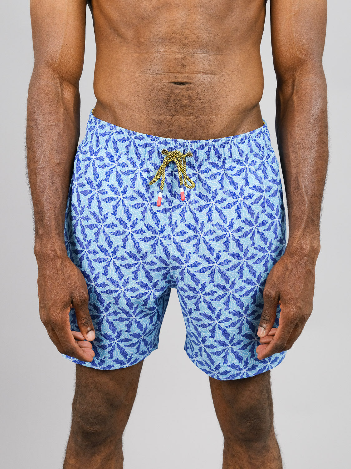 A person is wearing the Nalu Fast Dry 4-Way Stretch Swim Trunk by ID Menswear, featuring a blue leaf pattern on quick-dry fabric from recycled plastic bottles. The shorts have a yellow and green drawstring and are captured against a plain background, framing the person from neck to mid-thigh.