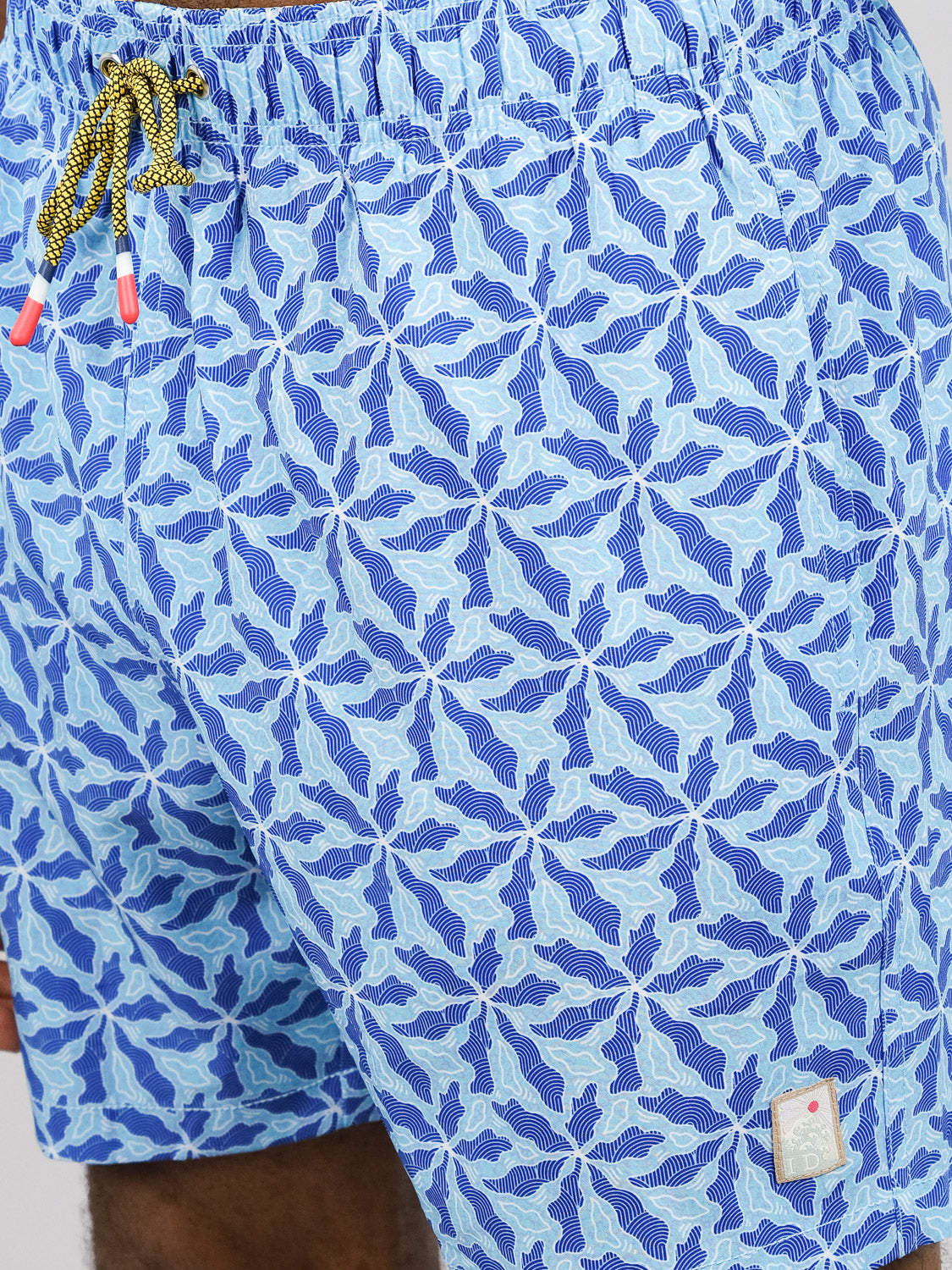 The Nalu Fast Dry 4-Way Stretch Swim Trunk by ID Menswear features a blue geometric leaf print, yellow drawstrings with red tips, a white hem patch, and is crafted from quick-dry, 4-way stretch fabric for maximum comfort.