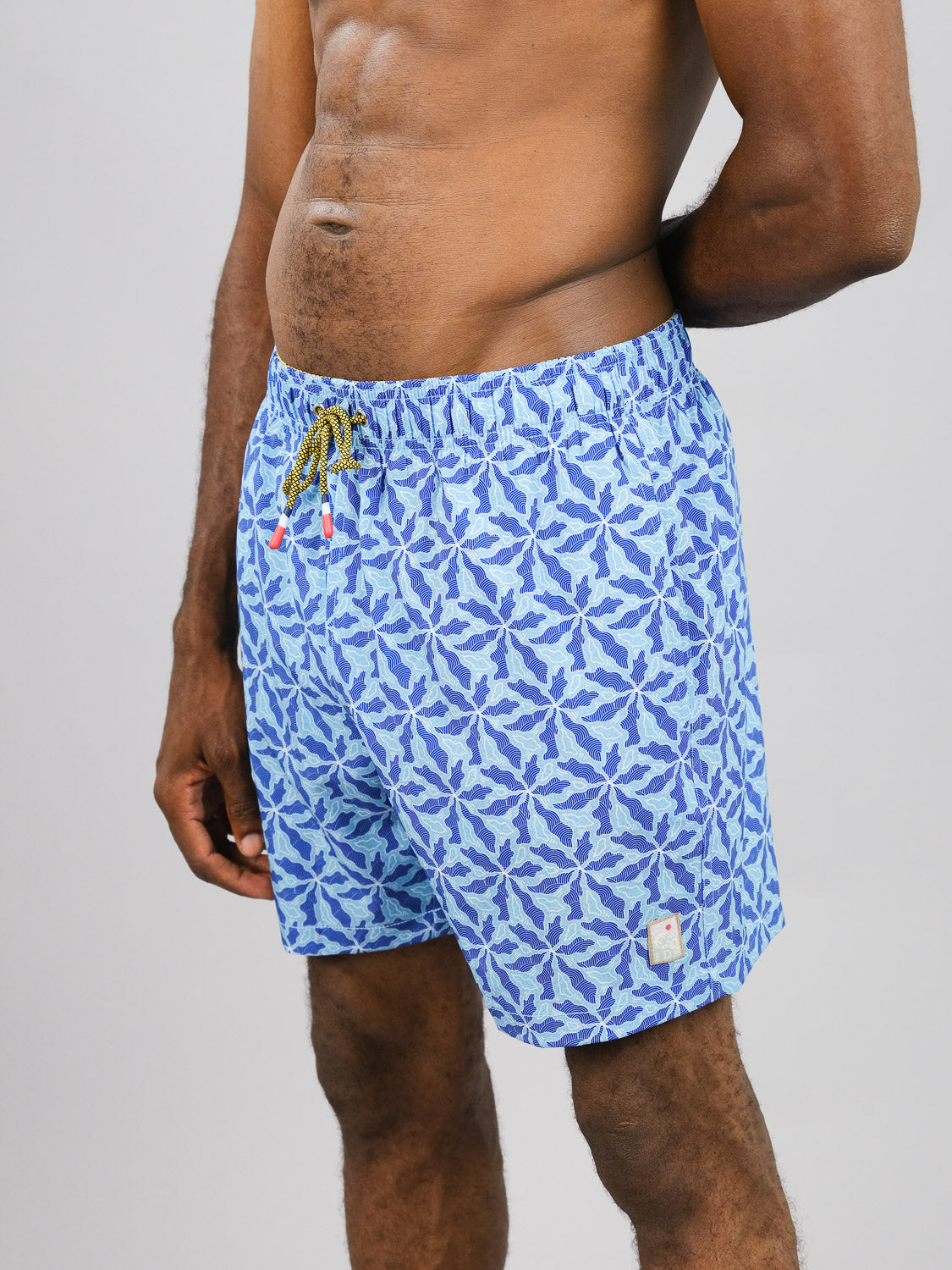 A person stands against a plain light gray background wearing ID Menswears Nalu Fast Dry 4-Way Stretch Swim Trunk. These blue swim trunks feature a white leaf pattern, are made from recycled plastic bottles, and have a drawstring waist for comfort.