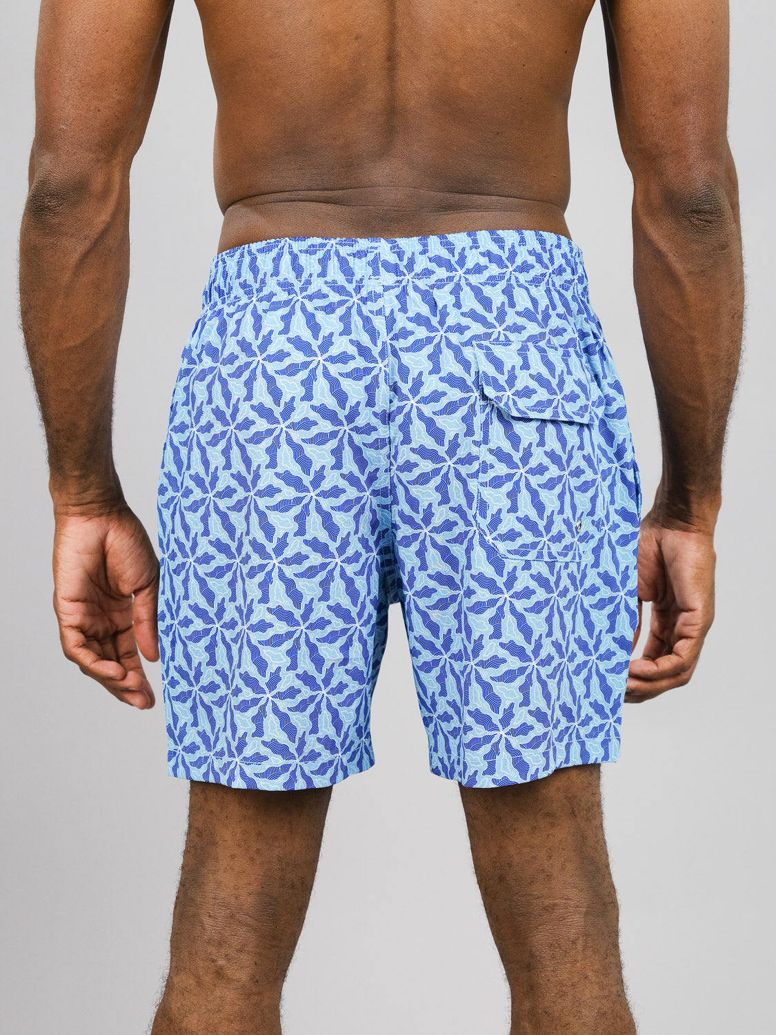 A person showcases ID Menswears Nalu Fast Dry 4-Way Stretch Swim Trunk, featuring blue and white abstract patterns on recycled plastic fabric. The vibrant shorts highlight quick-dry technology and maximum comfort against a gray background.