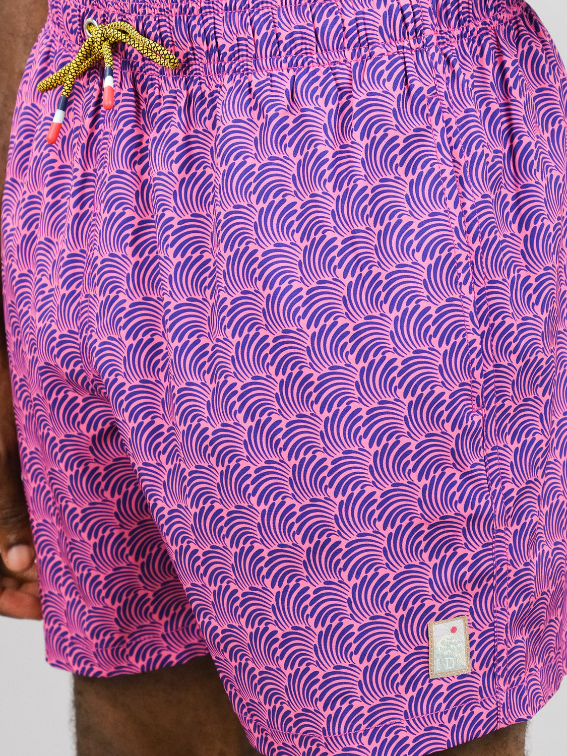 Close-up of a person in ID Menswear’s Nazare Fast Dry 4-Way Stretch Swim Trunk—pink with blue wave pattern, yellow-brown drawstring, small white hem label, and made from recycled plastic bottles.