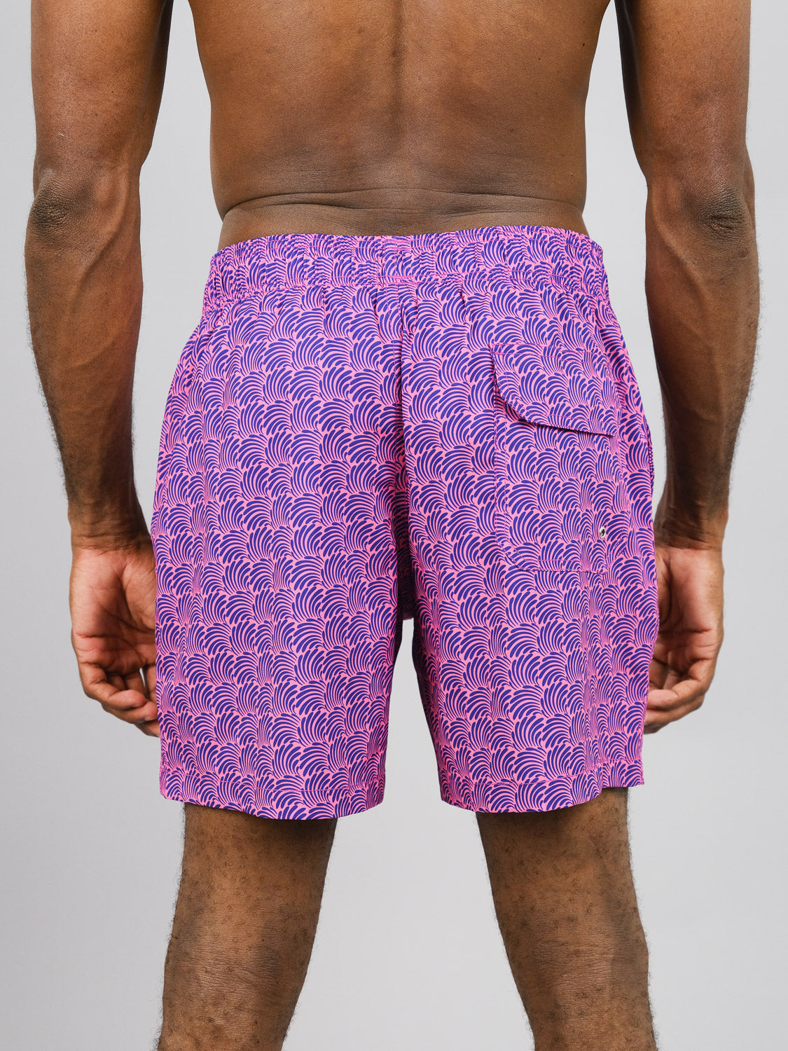 A person wearing Nazare Fast Dry 4-Way Stretch Swim Trunk by ID Menswear, featuring pink shorts with a purple wave pattern made from recycled plastic, is viewed from the back. These quick-dry shorts have an elastic waistband and a right-side pocket, set against a plain gray background.