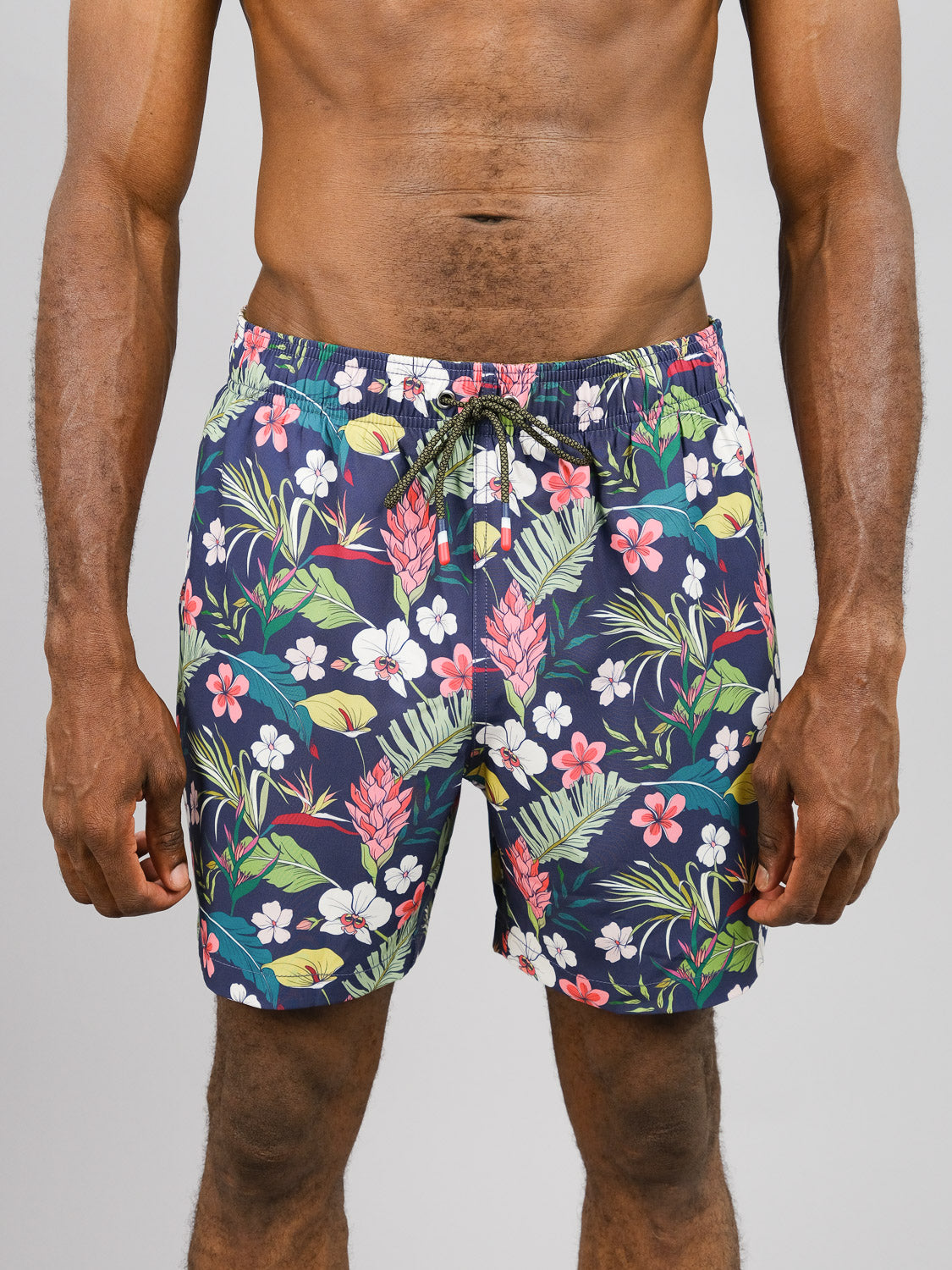 Someone is wearing ID Menswears Kipukai Fast Dry 4-Way Stretch Swim Trunk in a blue tropical pattern with pink, red, and white flowers as well as lush green leaves. Made from recycled plastic bottles, these shorts have a drawstring waist. The backdrop is plain light gray.