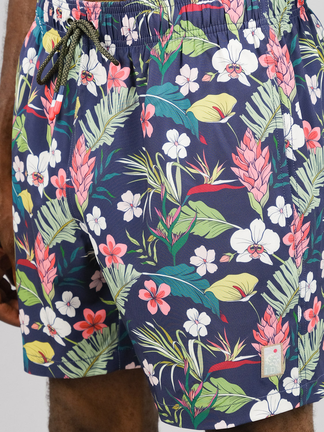 Close-up of a person wearing the ID Menswear Kipukai Fast Dry 4-Way Stretch Swim Trunk, featuring a navy base with vibrant pink, white, green, and red tropical floral print and a drawstring waist in quick-dry fabric.