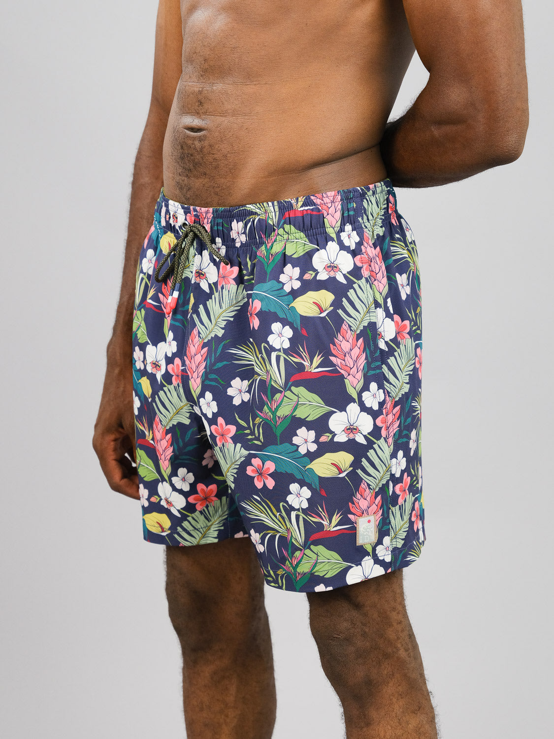 A person in Kipukai Fast Dry 4-Way Stretch Swim Trunk by ID Menswear stands against a plain gray background. Made from recycled plastic bottles, these vibrant shorts feature pink, white, and green tropical flowers and leaves for a stylishly sustainable beach vibe.