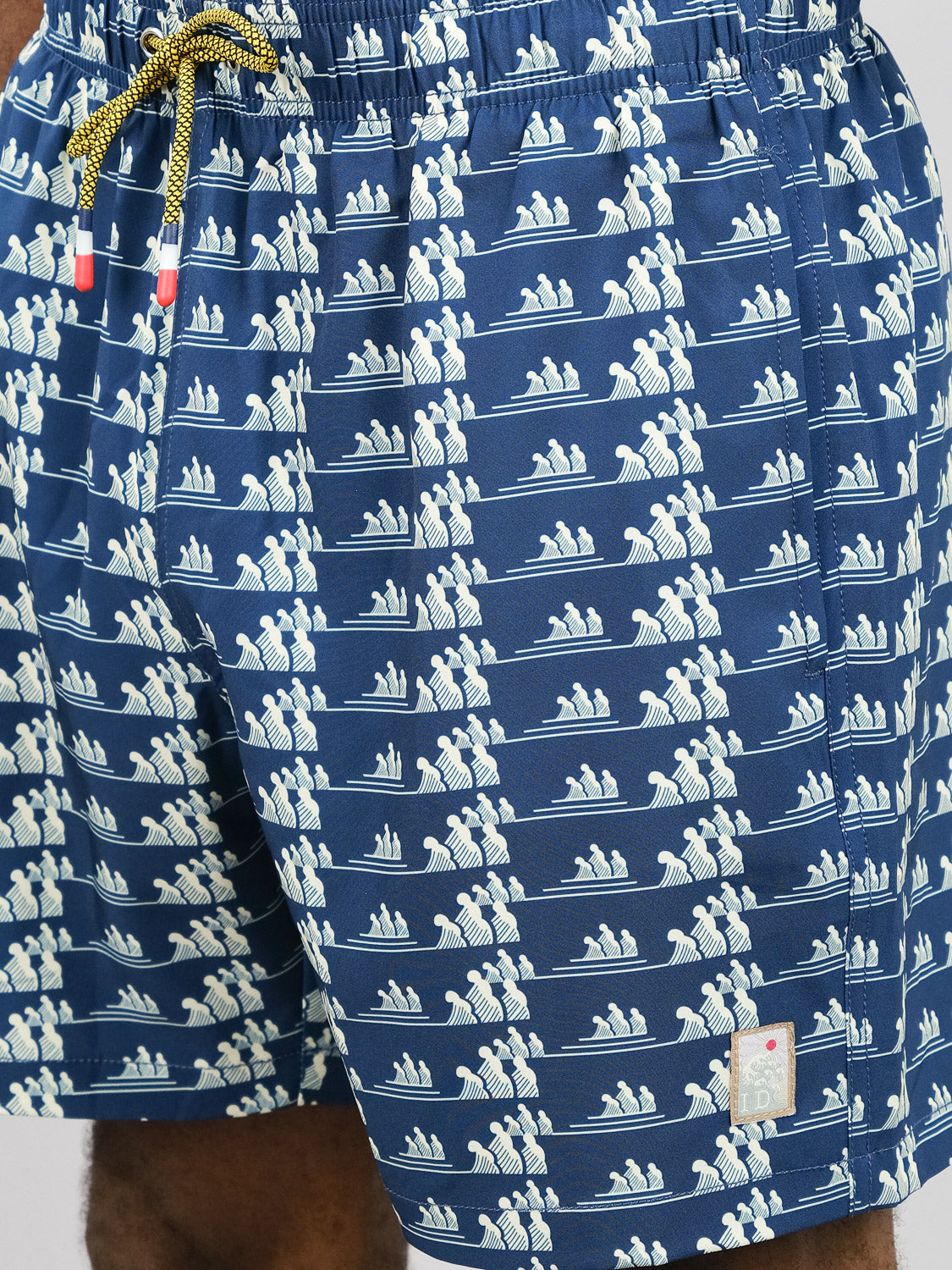 A close-up of the Tsunami Fast Dry 4-Way Stretch Swim Trunk by ID Menswear in blue, featuring a white rowing pattern, yellow drawstrings, quick-dry fabric, and a small square patch near the hem.