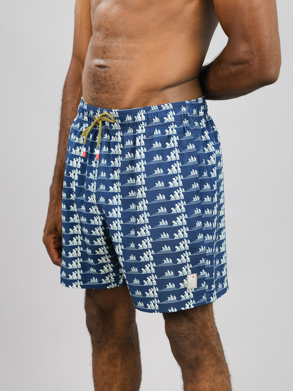 A shirtless individual stands against a gray background, wearing ID Menswears Tsunami Fast Dry 4-Way Stretch Swim Trunk. The eco-conscious blue shorts, featuring a white sailboat pattern and made from recycled plastic bottles, are secured by a yellow drawstring. Their left hand touches their right elbow.