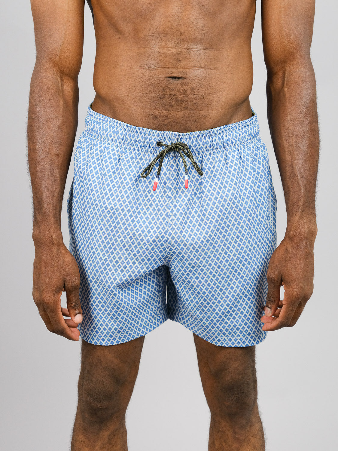 A person wears ID Menswears Stars Fast Dry 4-Way Stretch Swim Trunk, featuring a geometric design and made from recycled bottles, set against a plain gray background. Only their torso and legs are visible.