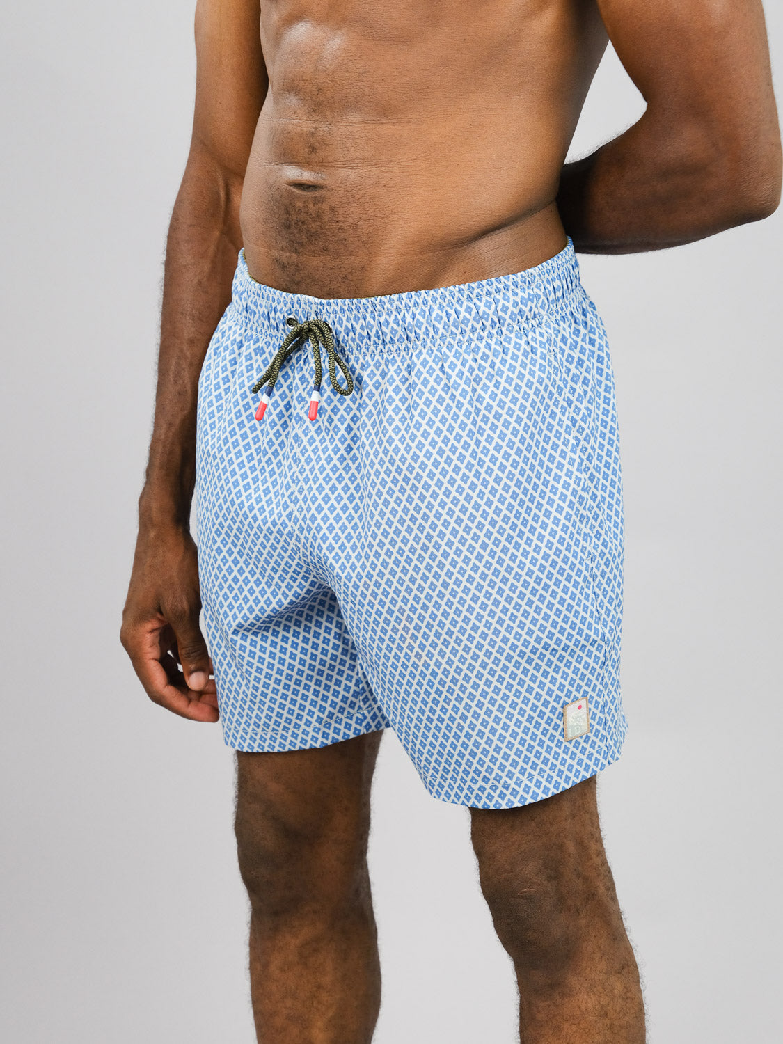 Against a gray background, a person wears ID Menswears Stars Fast Dry 4-Way Stretch Swim Trunk, crafted from recycled plastic bottles. The light blue shorts feature geometric patterns, a drawstring waist, and a logo patch on the lower left side for comfort and style.