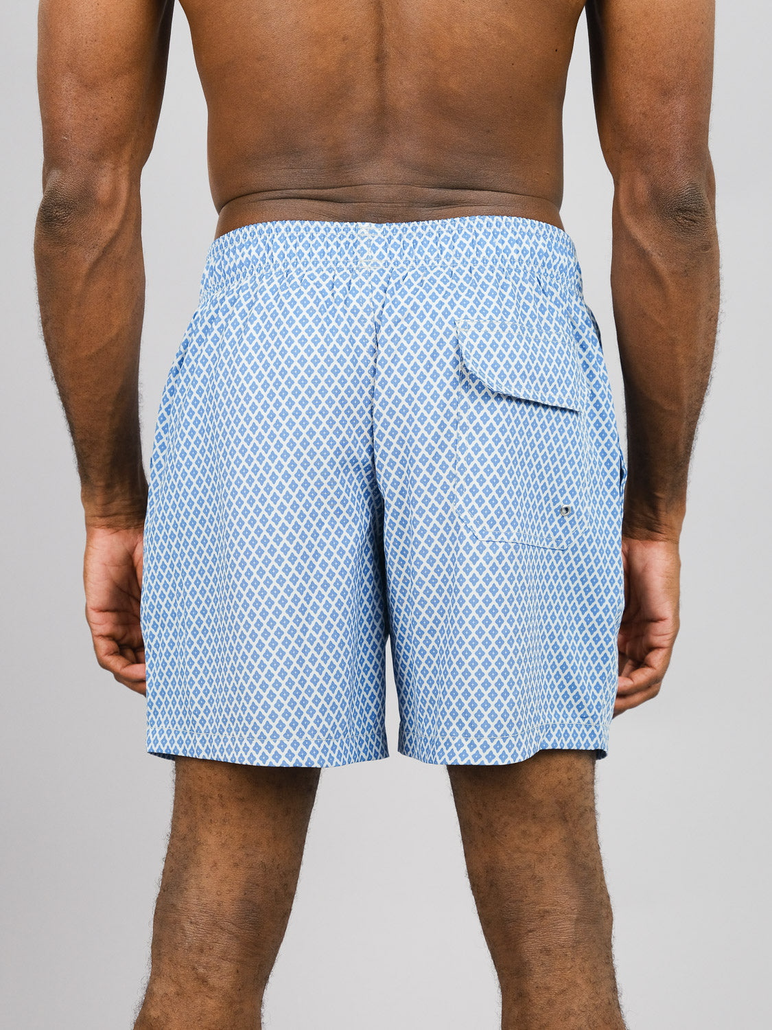 A man with medium to dark skin tone wears ID Menswears Stars Fast Dry 4-Way Stretch Swim Trunk, light blue and white, made from recycled bottles. Features include an elastic waistband and a back pocket. The background is plain light gray.
