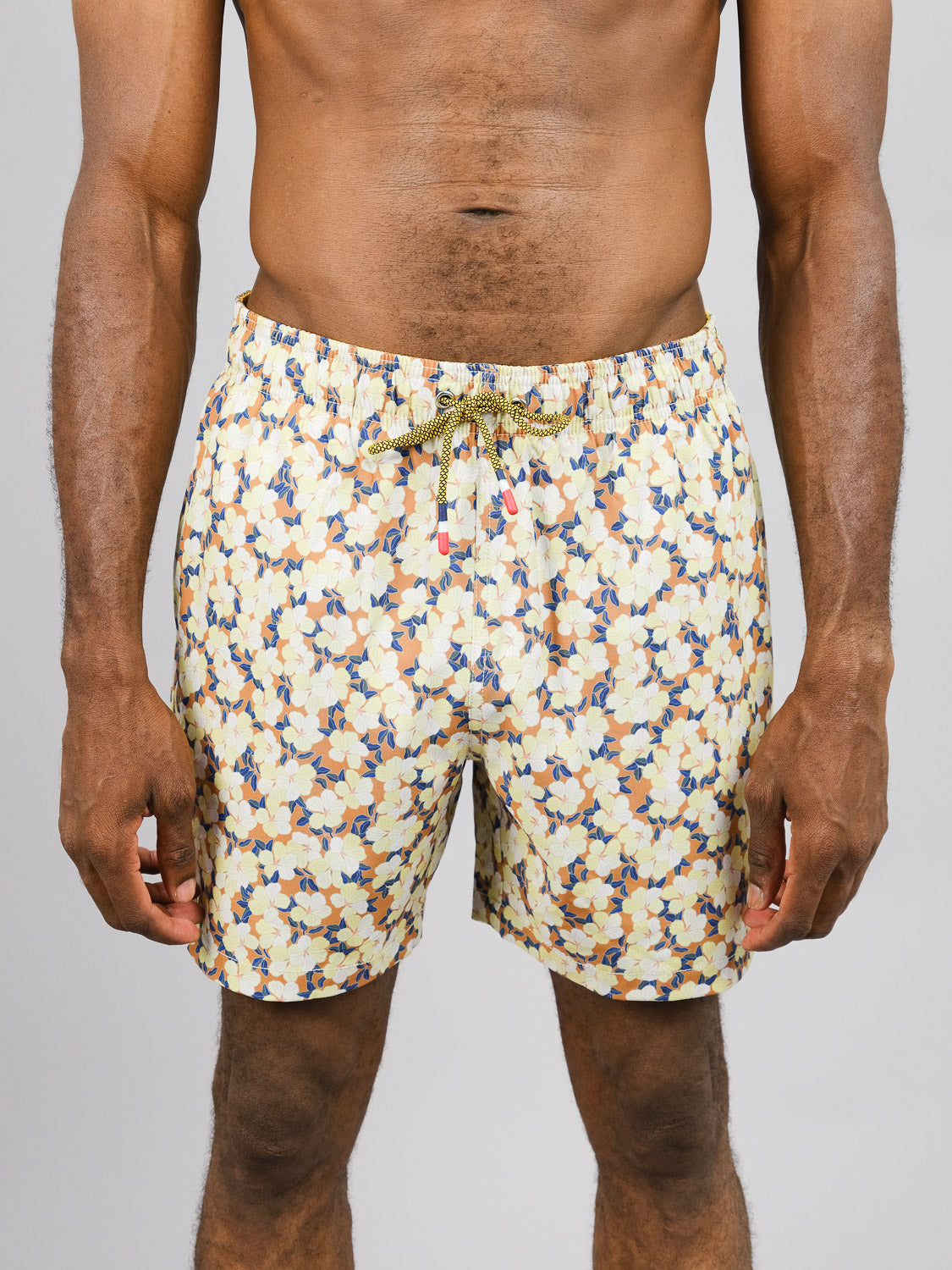 A person models Kokio Fast Dry 4-Way Stretch Swim Trunk by ID Menswear, crafted from recycled plastic bottles. The floral-patterned shorts feature vibrant yellow, blue, and orange hues against a plain background, showcasing their eco-friendly design.