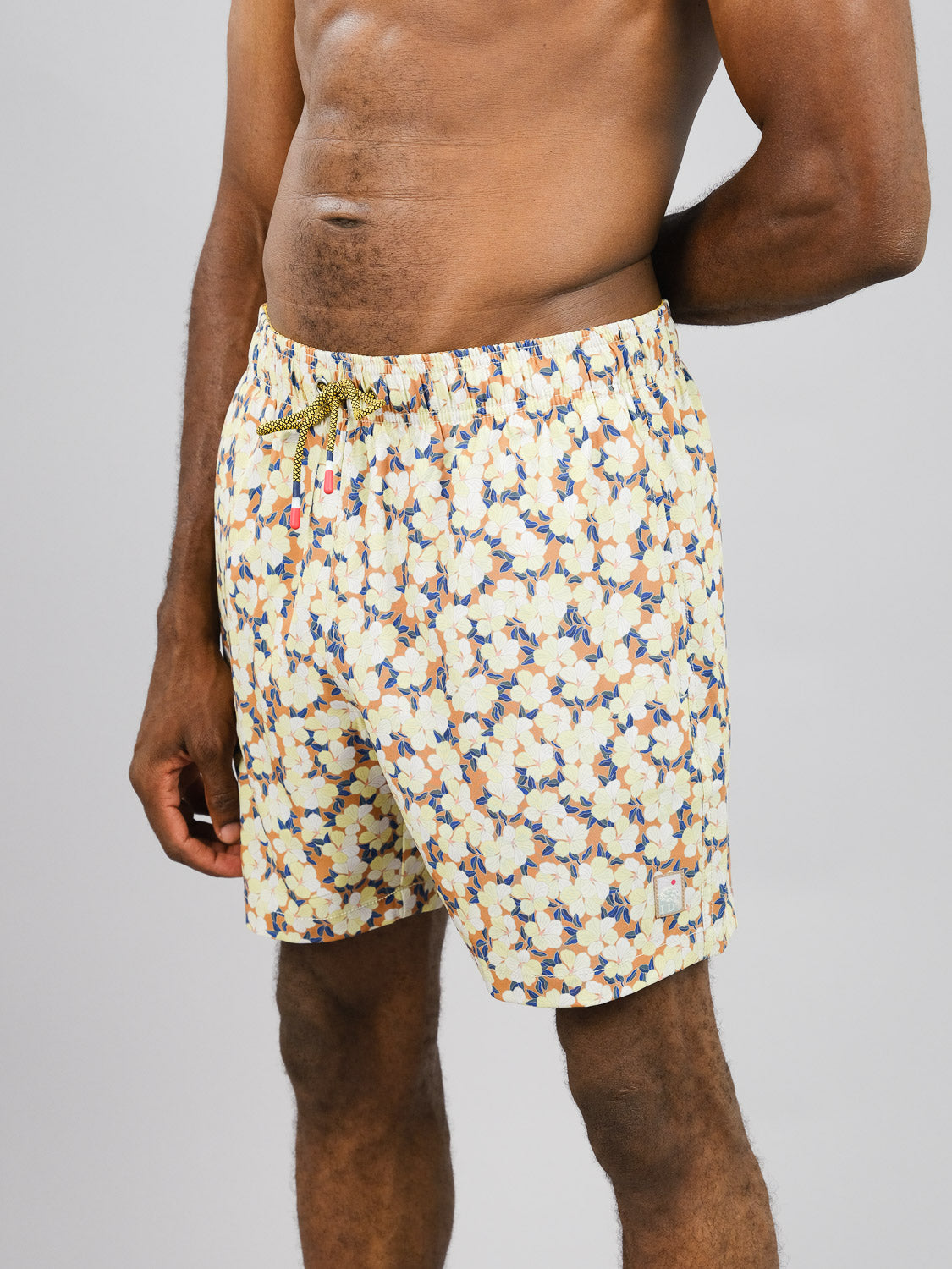 A person poses against a plain gray backdrop wearing ID Menswears Kokio Fast Dry 4-Way Stretch Swim Trunk, featuring a yellow, brown, and blue floral pattern made from recycled plastic bottles. The trunks have a drawstring waistband for comfort, with one hand resting casually on their hip.
