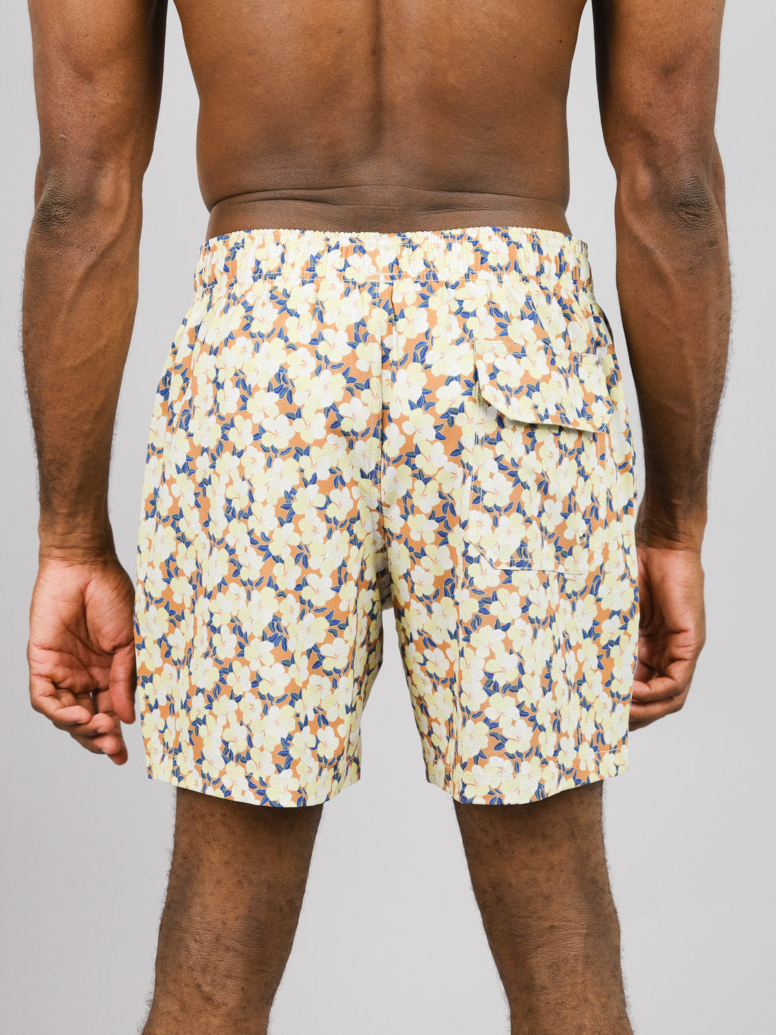 Someone is wearing Kokio Fast Dry 4-Way Stretch Swim Trunks from ID Menswear, featuring a floral pattern in light yellow, blue, and beige. Made from quick-dry fabric crafted from recycled plastic bottles, they include an elastic waistband and back pocket on a plain gray background for an eco-friendly design.