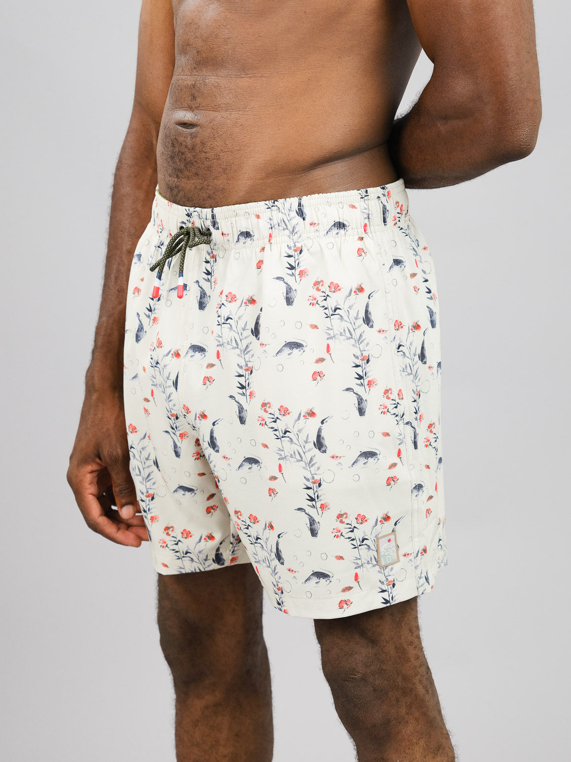A person is wearing ID Menswears Koi Fast Dry 4-Way Stretch Swim Trunk in cream, featuring a red and blue floral pattern. They stand against a plain gray background, visible from the waist up, with their right arm resting at their side.