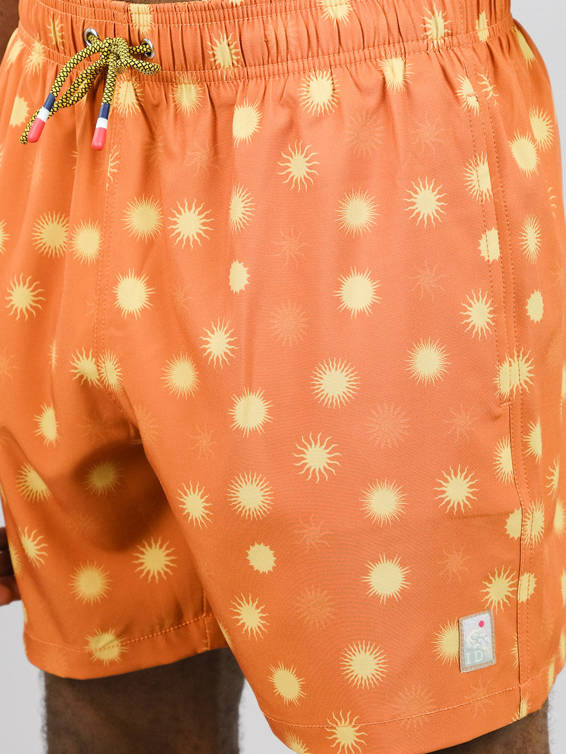 Close-up of a person in ID Menswear’s Sol Fast Dry 4-Way Stretch Swim Trunk, featuring an orange sunburst print, quick-dry fabric, green drawstring with colorful tips, and a logo tag near the hem.