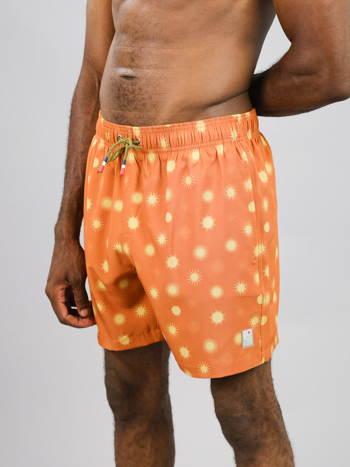 A person models the ID Menswear Sol Fast Dry 4-Way Stretch Swim Trunk, featuring an orange sunburst pattern. Made from recycled bottles, these trunks with an elastic waistband and multicolored drawstring stand out against a plain background, with the persons upper body bare and arm relaxed by their side.
