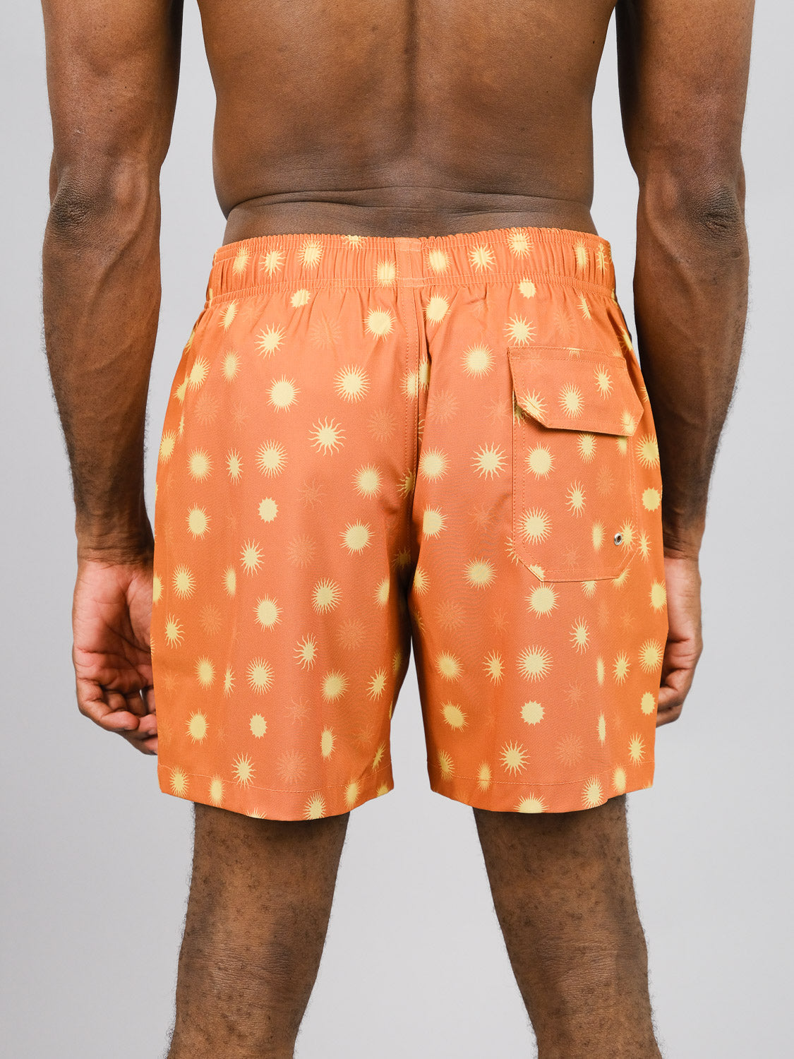 A person is seen from behind wearing ID Menswears Sol Fast Dry 4-Way Stretch Swim Trunk, featuring a yellow starburst pattern on orange and made from recycled plastic bottles. The shorts have a waistband and back pocket, providing stretch for comfort against a plain gray background.