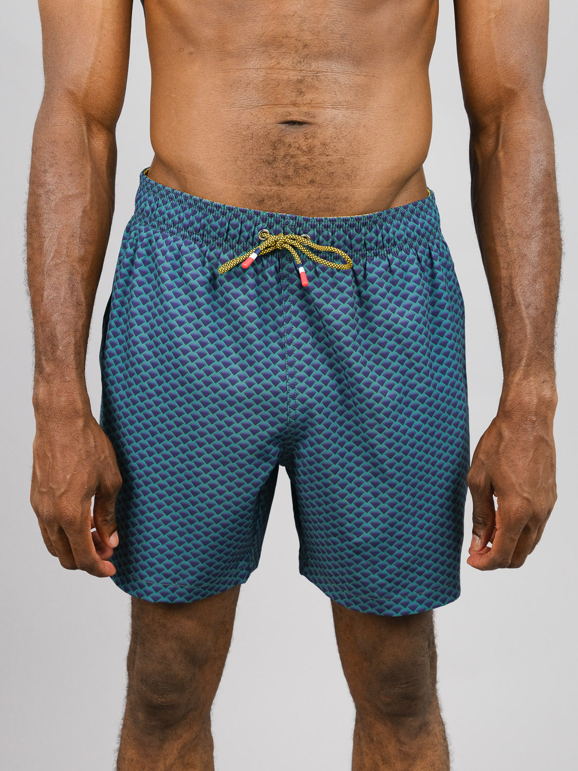 Against a plain light gray backdrop, a person wears ID Menswears Uroko Fast Dry 4-Way Stretch Swim Trunk in trendy blue and green patterns. Made from quick-dry fabric and partially recycled plastic bottles, these drawstring shorts offer both comfort and style.