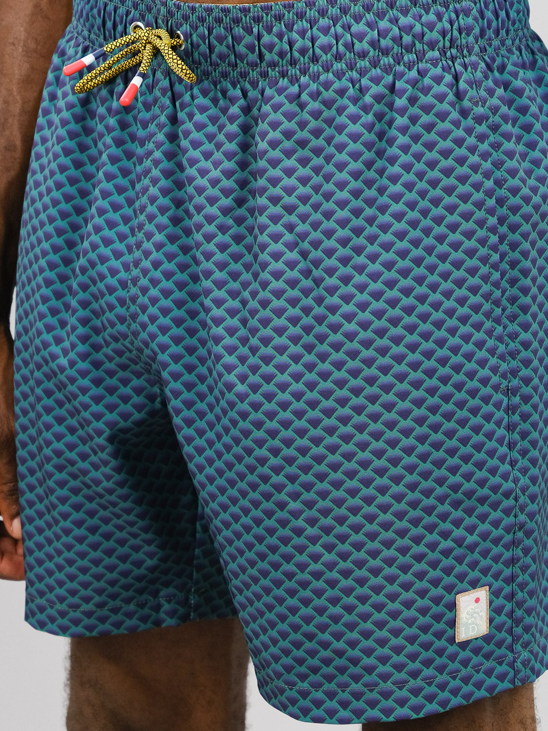 Close-up of a person in ID Menswears Uroko Fast Dry 4-Way Stretch Swim Trunk, featuring blue and green geometric patterns, yellow-black drawstring, small rectangular patch near the hem, and quick-dry fabric for all-day comfort.
