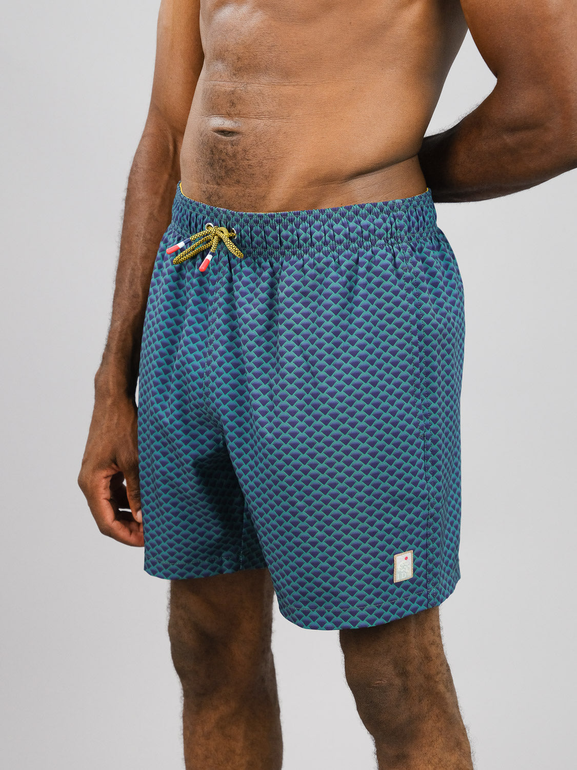 Against a plain backdrop, an individual models the Uroko Fast Dry 4-Way Stretch Swim Trunk by ID Menswear in blue with quick-dry fabric and drawstring. With a small tag near the hem, their stylish pose with bent left arm conveys effortless style and comfort.
