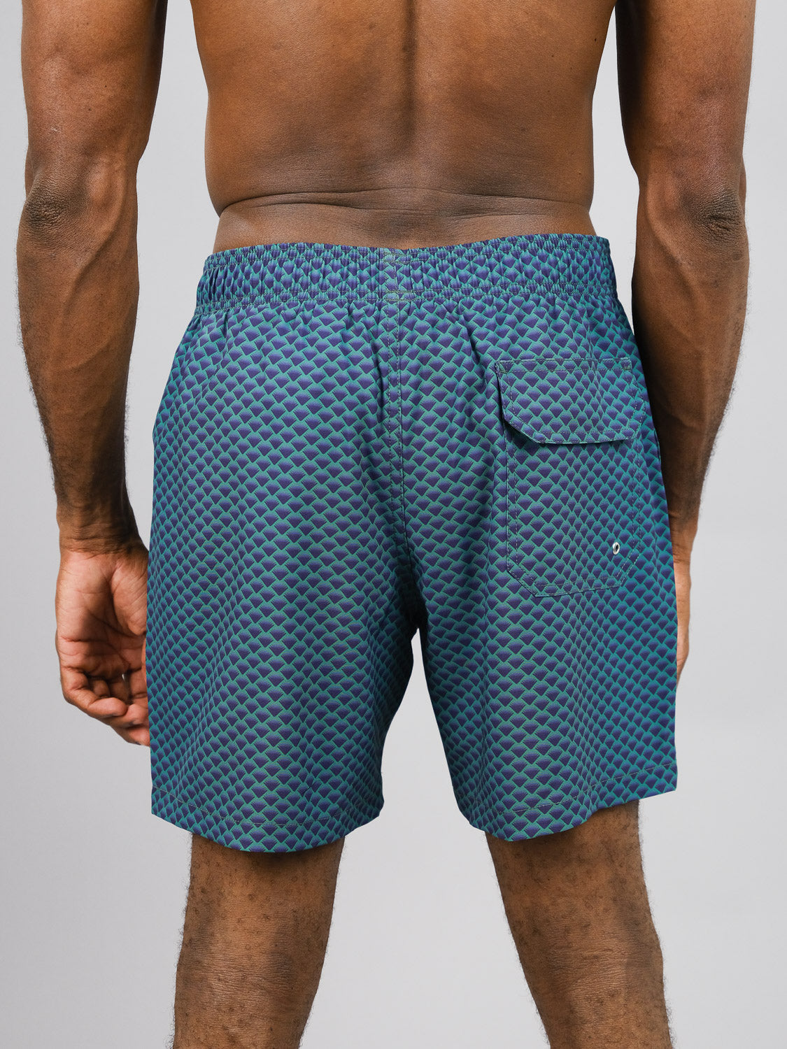 A person stands with their back to the camera wearing ID Menswears Uroko Fast Dry 4-Way Stretch Swim Trunk. These shorts, made from recycled plastic bottles, feature a geometric pattern, quick-dry fabric, an elastic waistband, and a back pocket against a plain light background.