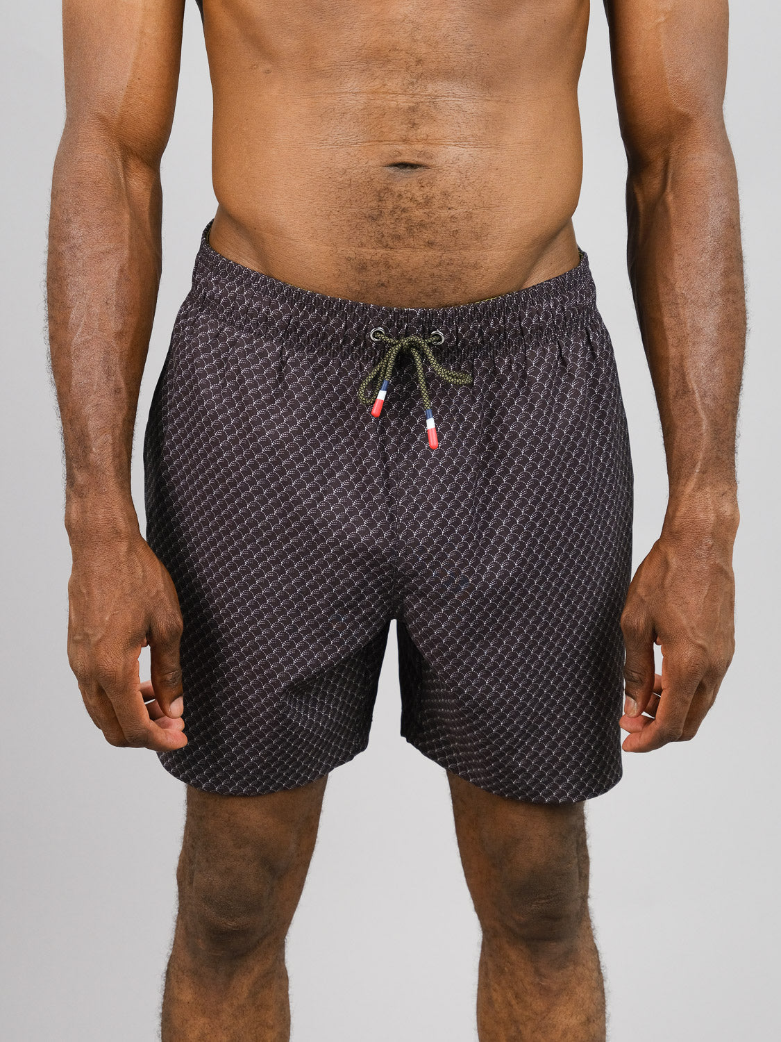 A person is wearing Broken Waves Fast Dry 4-Way Stretch Swim Trunk by ID Menswear, made from recycled plastic bottles. The black and white patterned shorts have quick-dry fabric and a drawstring with red and white tips. The view is from the shoulders to thighs against a light background.