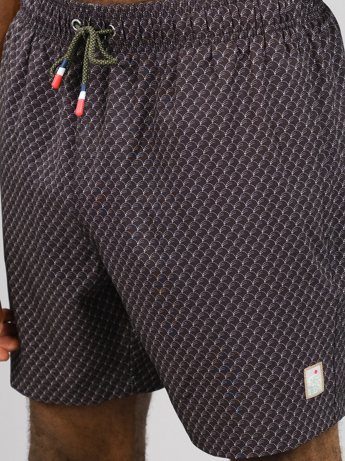 A close-up of someone wearing ID Menswear’s Broken Waves Fast Dry 4-Way Stretch Swim Trunk, featuring a dark geometric pattern, elastic waistband with green drawstrings, small white hem patch, and quick-dry fabric for all-day comfort.