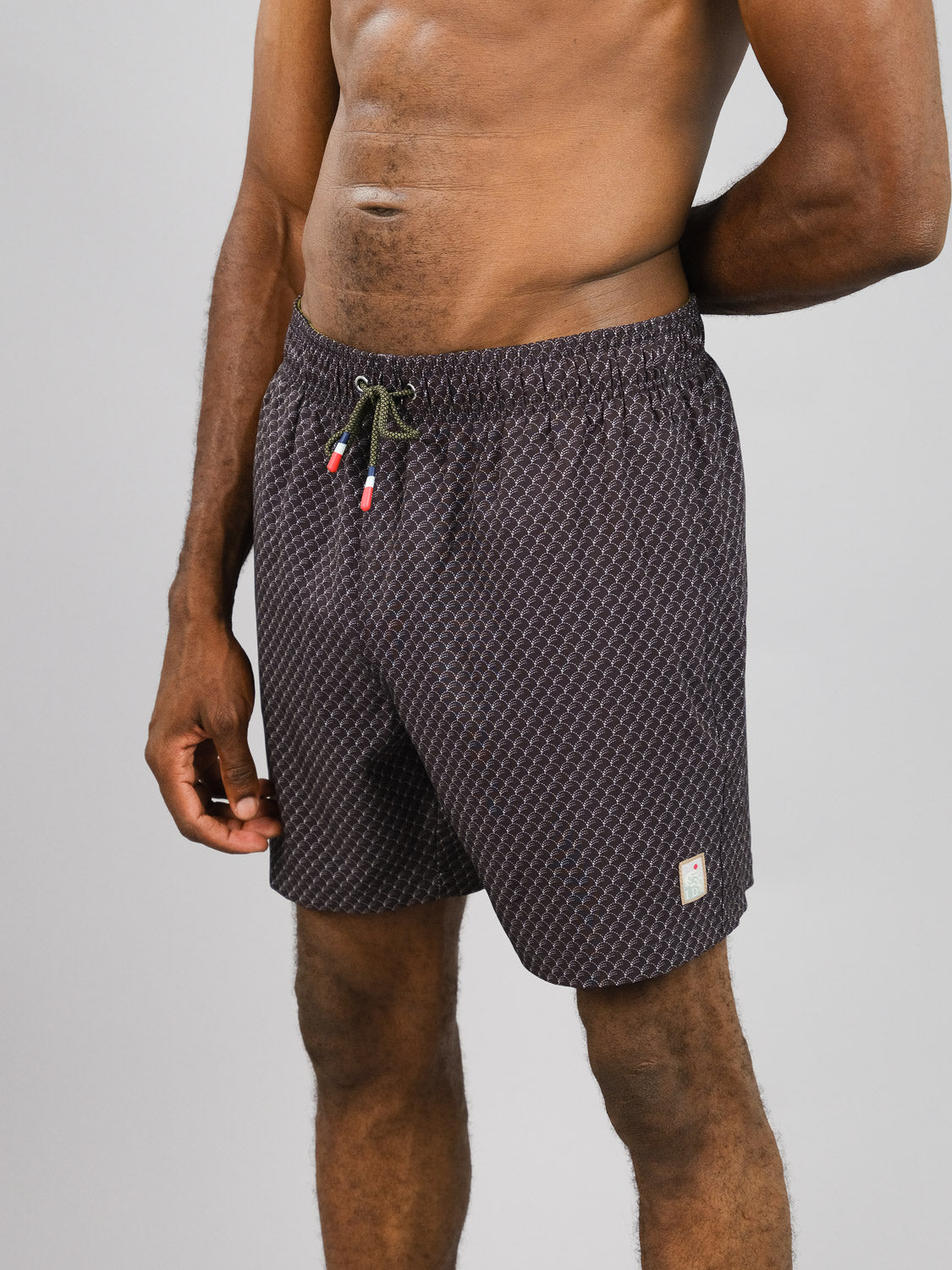 A shirtless person in Broken Waves Fast Dry 4-Way Stretch Swim Trunk by ID Menswear stands against a plain background. Made from recycled plastic bottles, these dark patterned shorts feature a drawstring and quick-dry fabric. Their left hand is partially visible at their side.