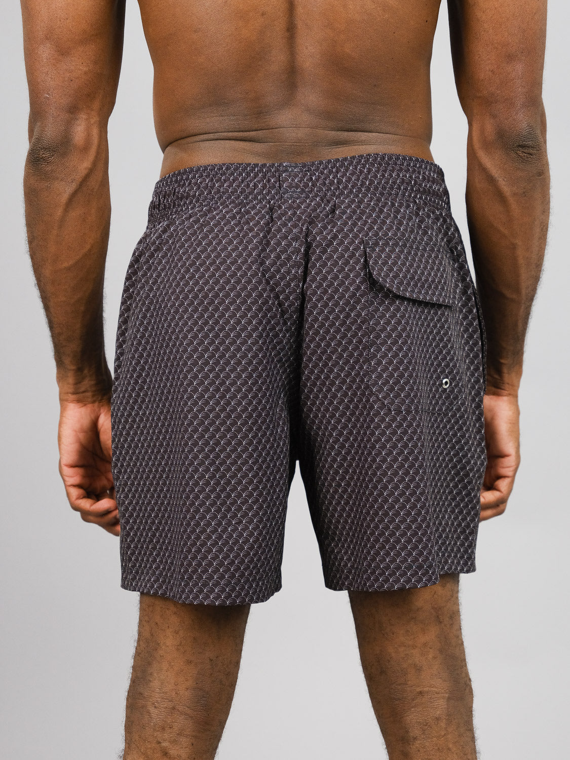 A person is seen from behind wearing ID Menswears Broken Waves Fast Dry 4-Way Stretch Swim Trunk, featuring patterned dark fabric, an elastic waistband, and a small back pocket on the right. The shorts combine comfort and functionality against a plain gray backdrop.