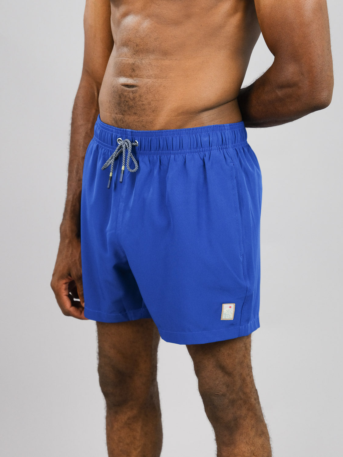 A person models Oyogu Quick Dry 4 Way Stretch Swim Trunk by ID Menswear, standing shirtless against a gray background. The bright blue recycled fabric shorts, made from plastic bottles, feature 4-way stretch and have a small patch near the hem; one arm rests by their side.