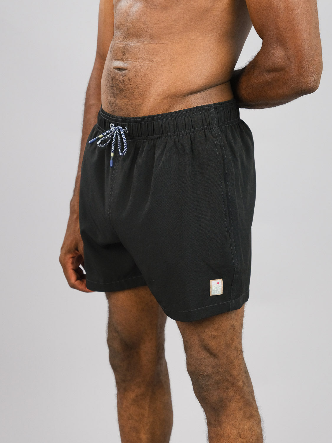 Someone stands against a plain background wearing Oyogu Quick Dry 4 Way Stretch Swim Trunk by ID Menswear, black with a blue drawstring and crafted from recycled plastic. The quick-dry shorts feature a small tag near the hem, while their upper body is partially out of view.
